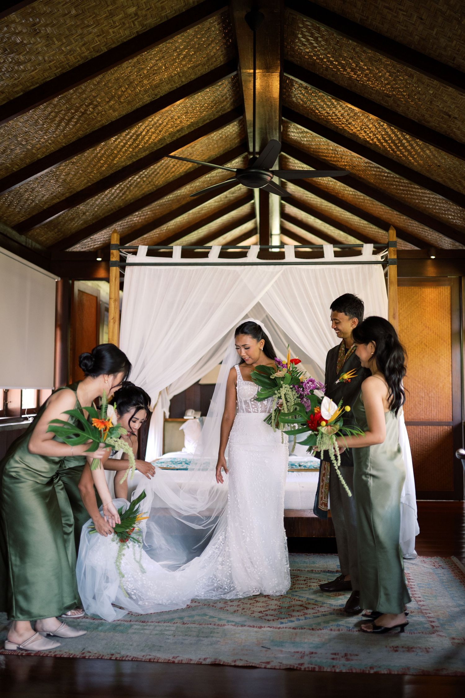 Bridesmaids in green satin dresses help bride prepare in elegant thatched-roof tropical villa.