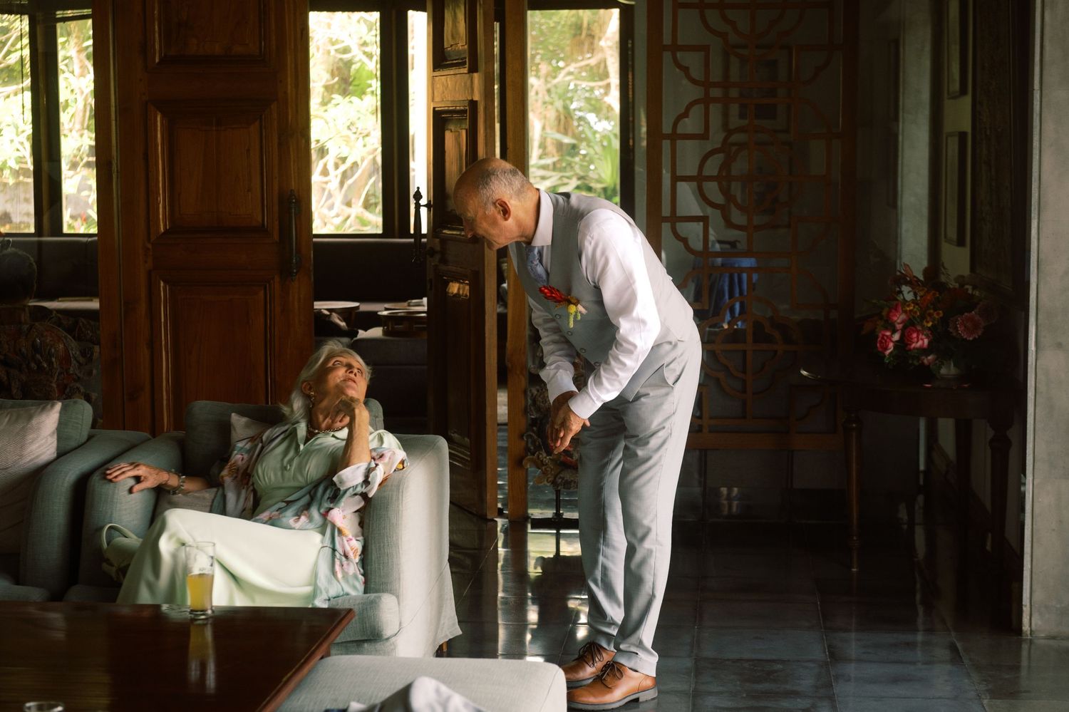 Groom assists seated guest in elegant villa interior during wedding day preparations.