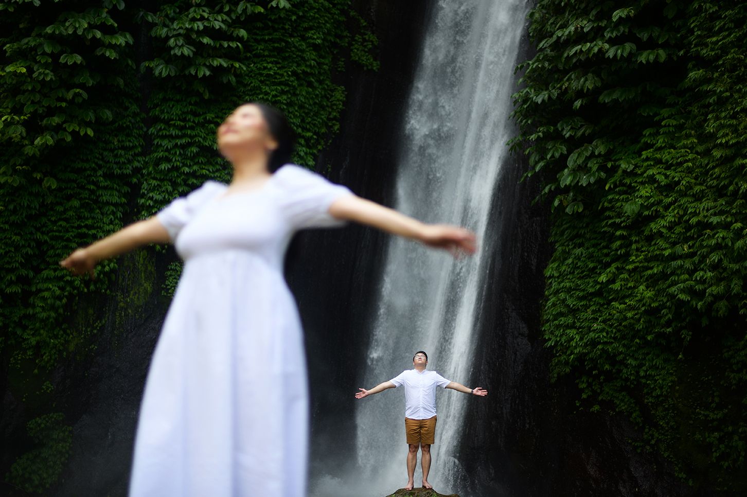 Woman in white dress and man pose with arms outstretched before a lush green waterfall backdrop.