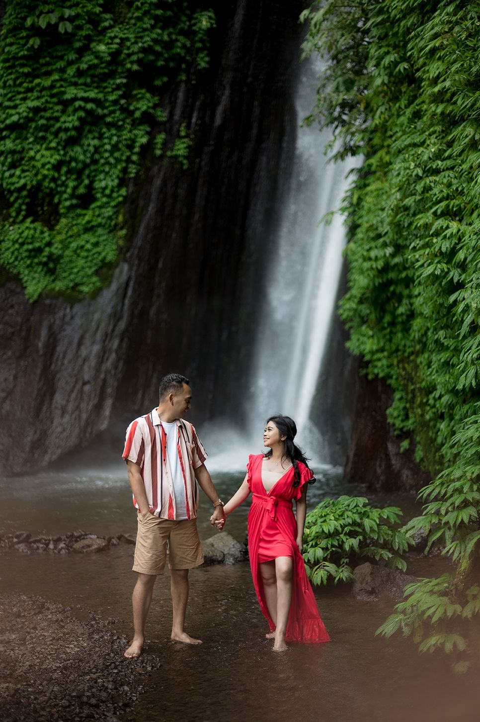 Couple holding hands barefoot by a lush tropical waterfall, woman in red dress, man in striped shirt and khaki shorts.