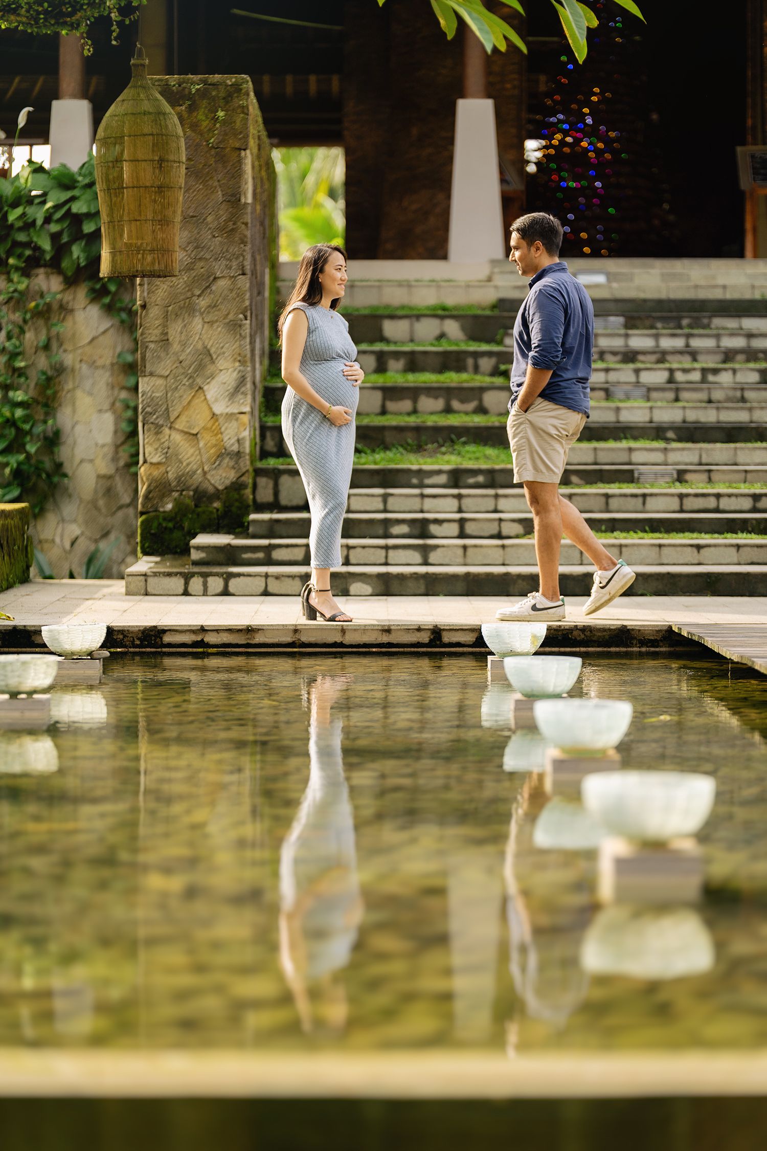 Pregnant woman and man standing by a reflective pool in a lush tropical garden setting Maya Ubud Resort and Spa in Bali.