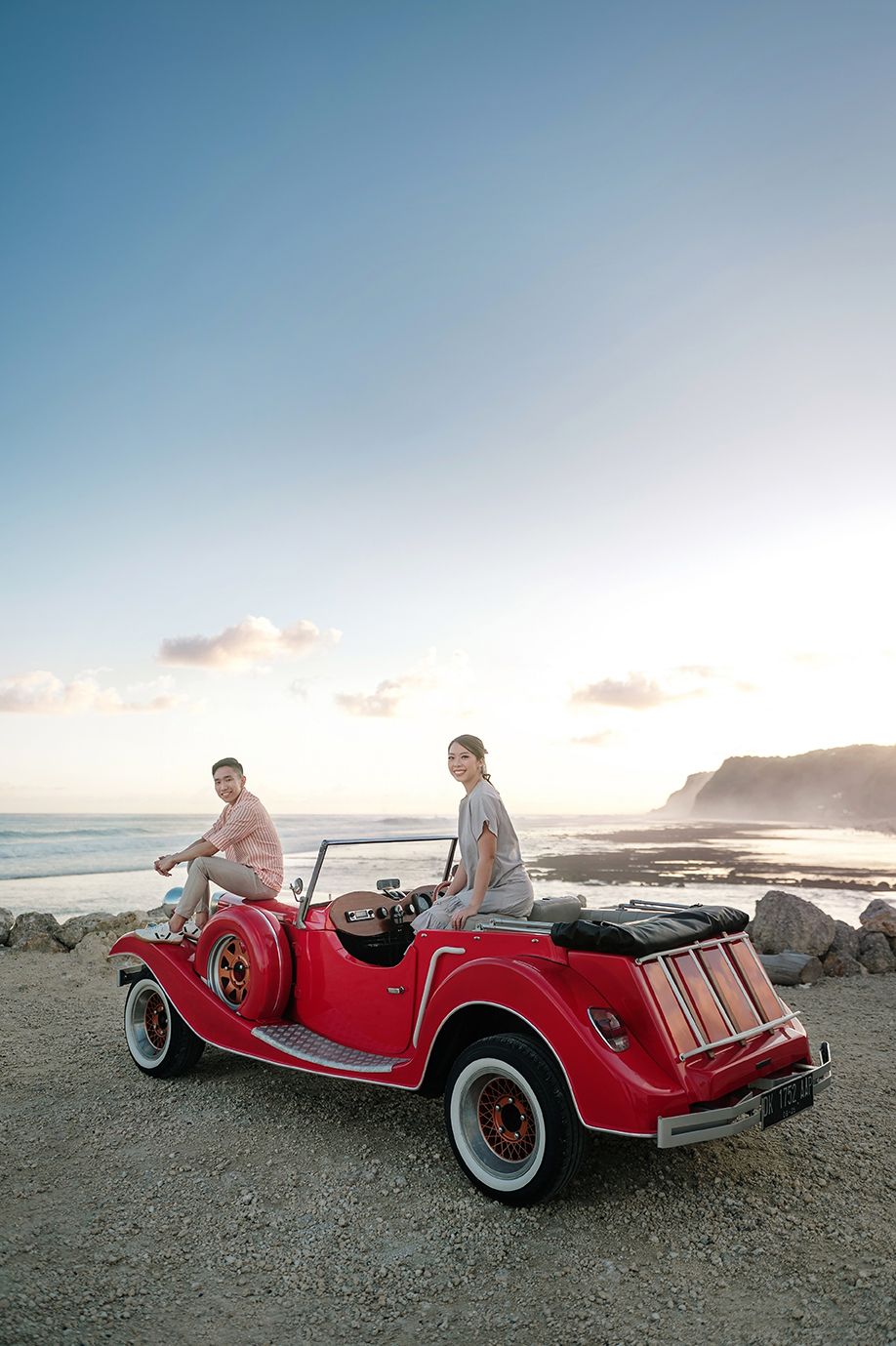 Couple posing with a classic red vintage convertible car on a rocky beach at sunset.