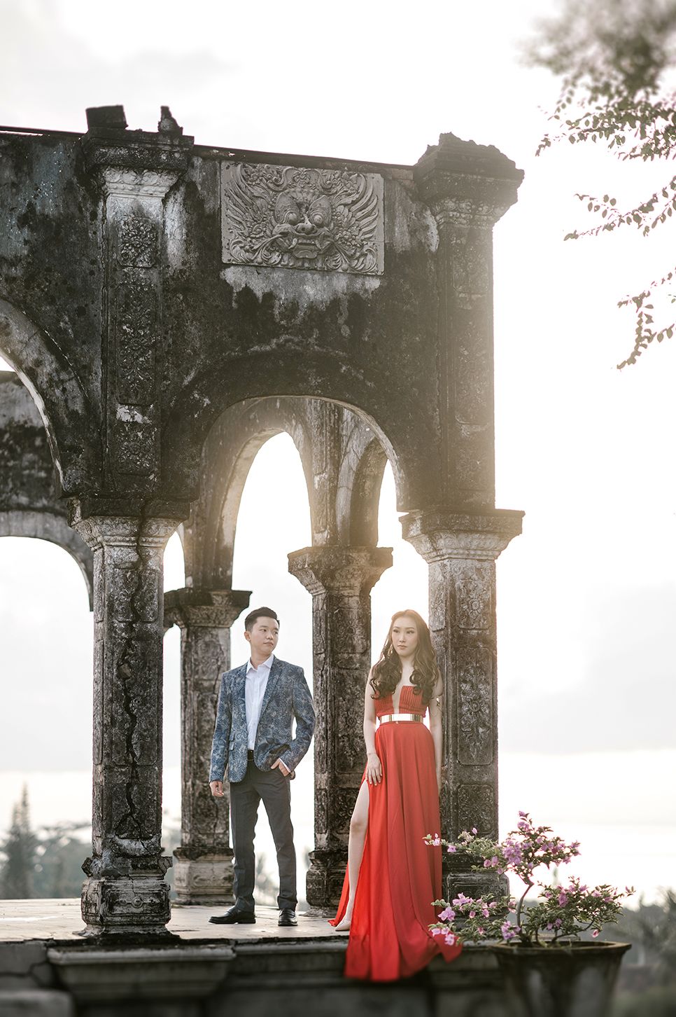 Couple posing at ancient stone archway ruins, woman in red gown, man in blue suit, dramatic backlight.