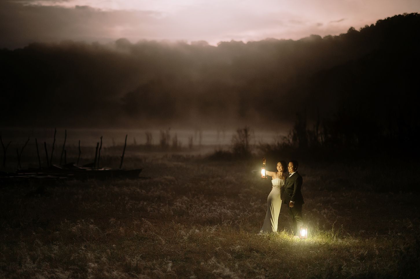 Couple holding glowing lanterns in a misty field at dusk with foggy hills and dramatic sky backdrop.