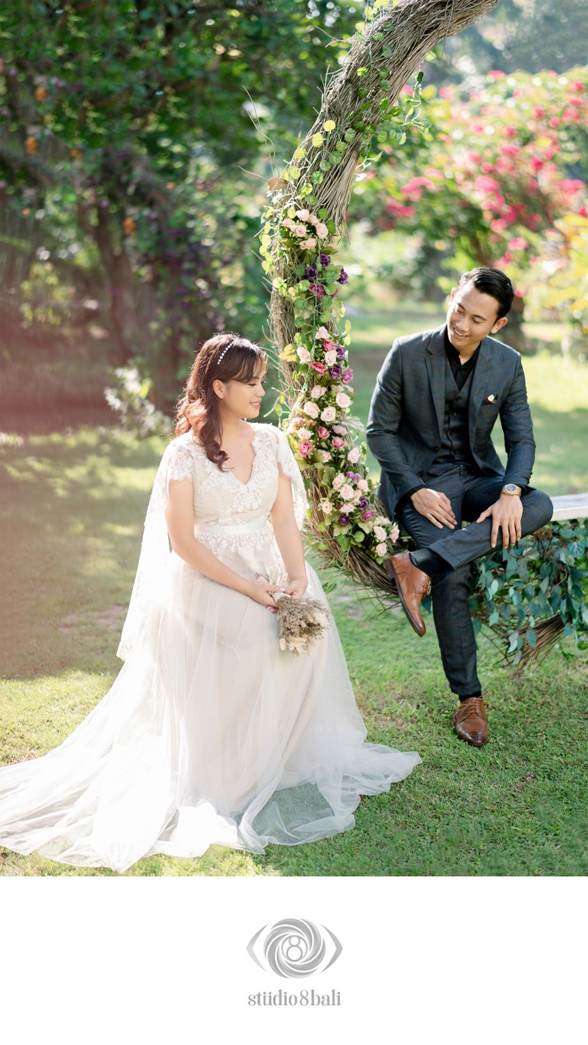 Couple in wedding attire posing in a lush garden, bride in white gown holding bouquet, groom in dark suit on floral swing.