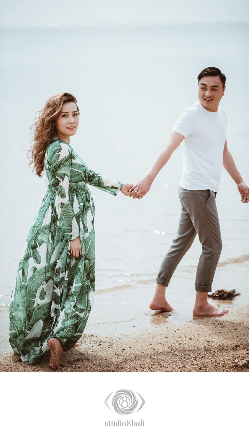 Couple holding hands on a sandy beach, woman in green floral maxi dress, man in white tee and gray pants.