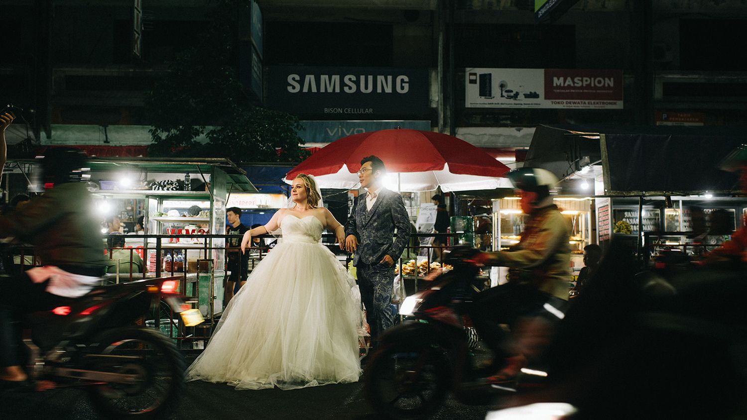 Bride in white gown and groom in suit pose on busy night street with Samsung sign and motorbikes in urban Asia.