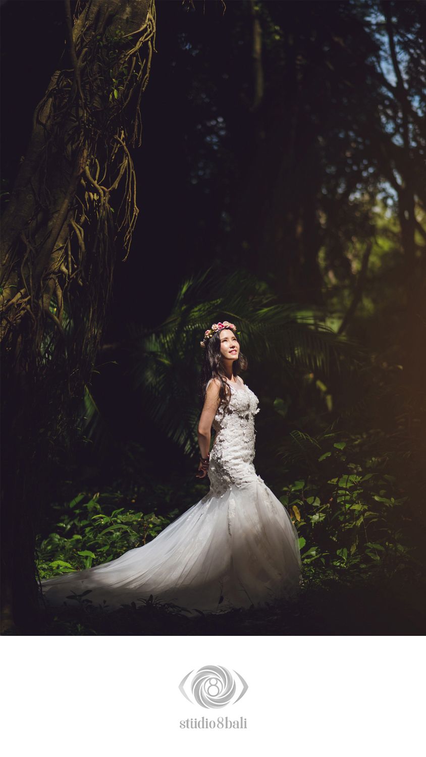 Bride in white mermaid gown with floral crown poses in a lush tropical forest, ethereal light illuminating her elegant silhouette.
