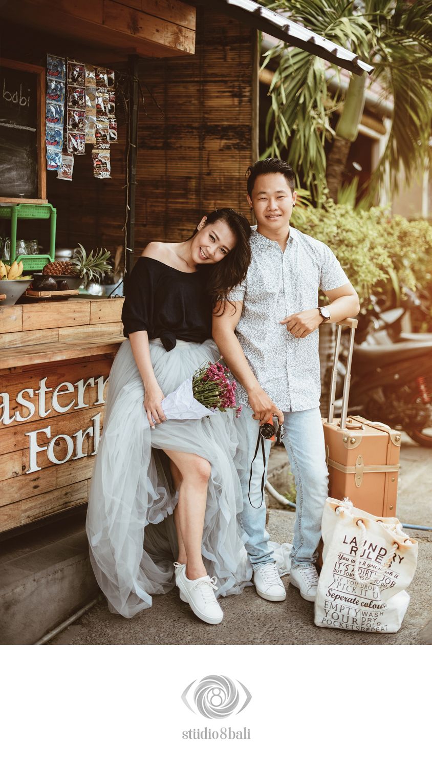 A couple poses together outside a rustic wooden cafe, the woman in a black top and tulle skirt holding flowers.