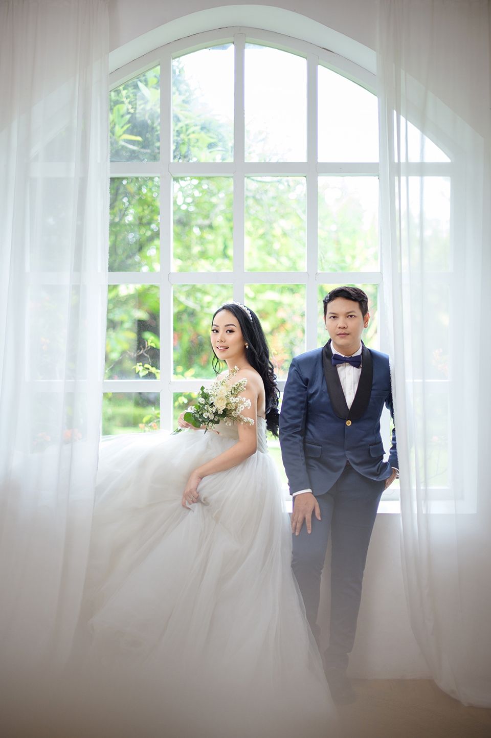 Elegant Asian couple in wedding attire posing by a large arched window, bride in white gown holding bouquet, groom in navy suit.
