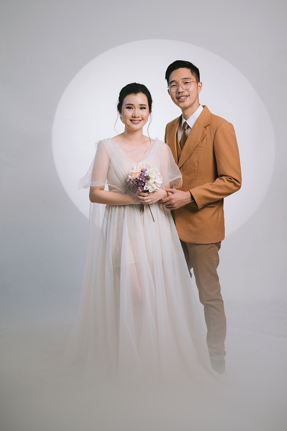 A couple poses for wedding photos, bride in white gown with bouquet, groom in tan suit, studio background.