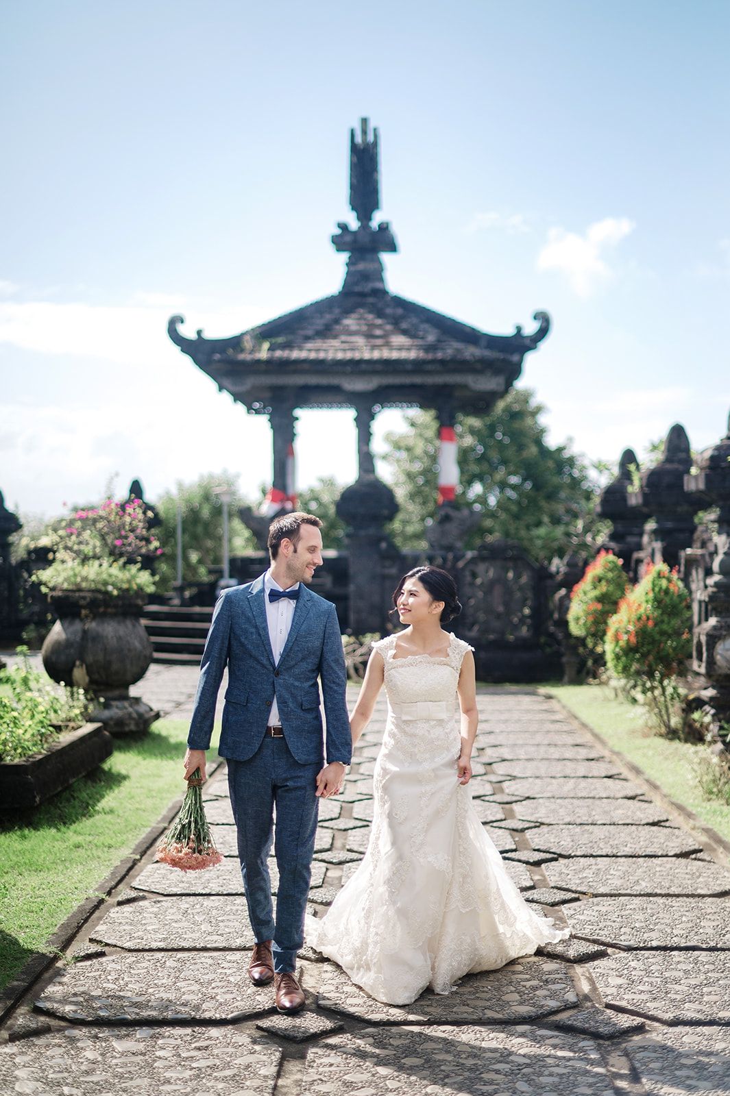 Bride and groom walk hand-in-hand along a stone path at a Balinese temple, surrounded by lush gardens.
