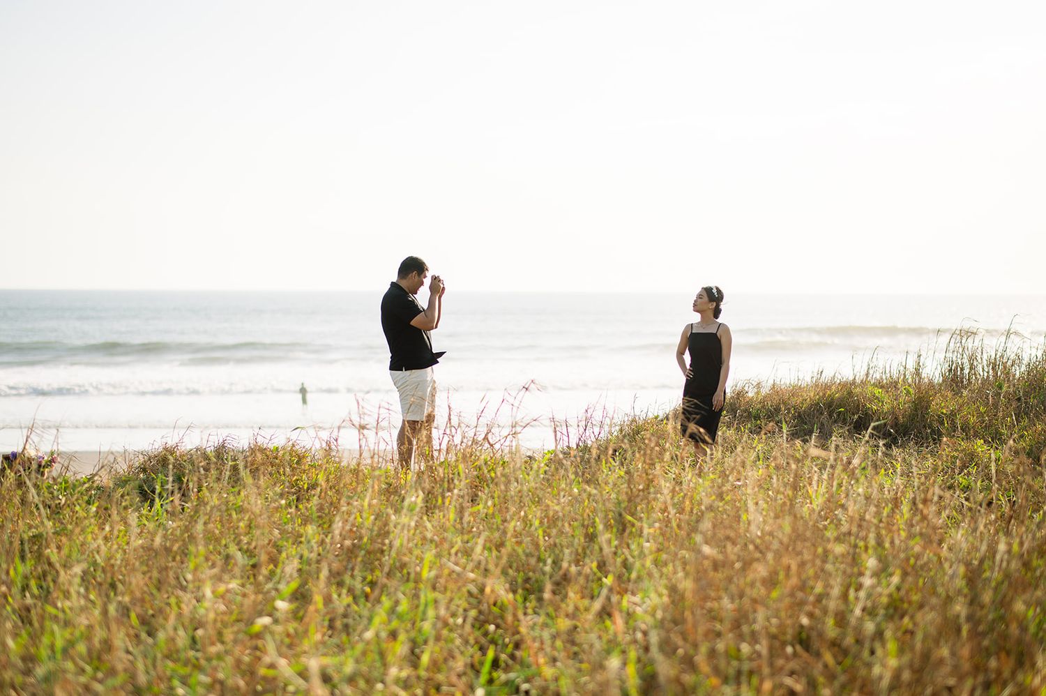 Man photographing woman on grassy coastal bluff with ocean waves in background on sunny day.