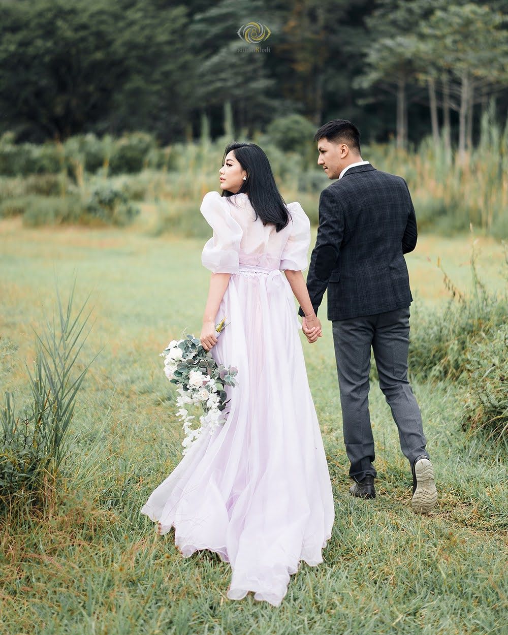 Couple holding hands walking through a lush green meadow, bride in pink gown with bouquet, groom in dark suit.