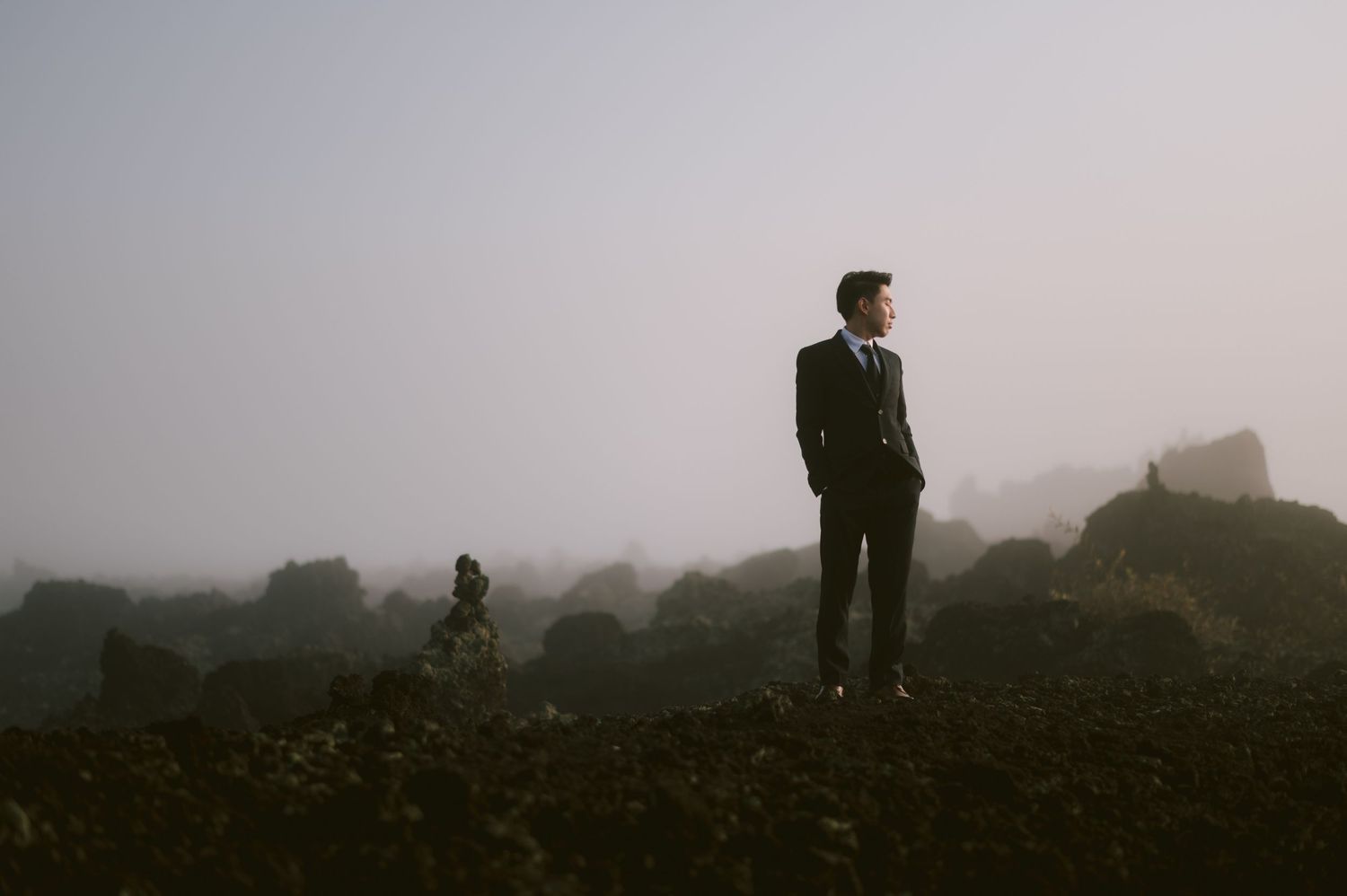A man in a dark suit stands confidently on rocky terrain surrounded by misty fog in a dramatic outdoor setting.