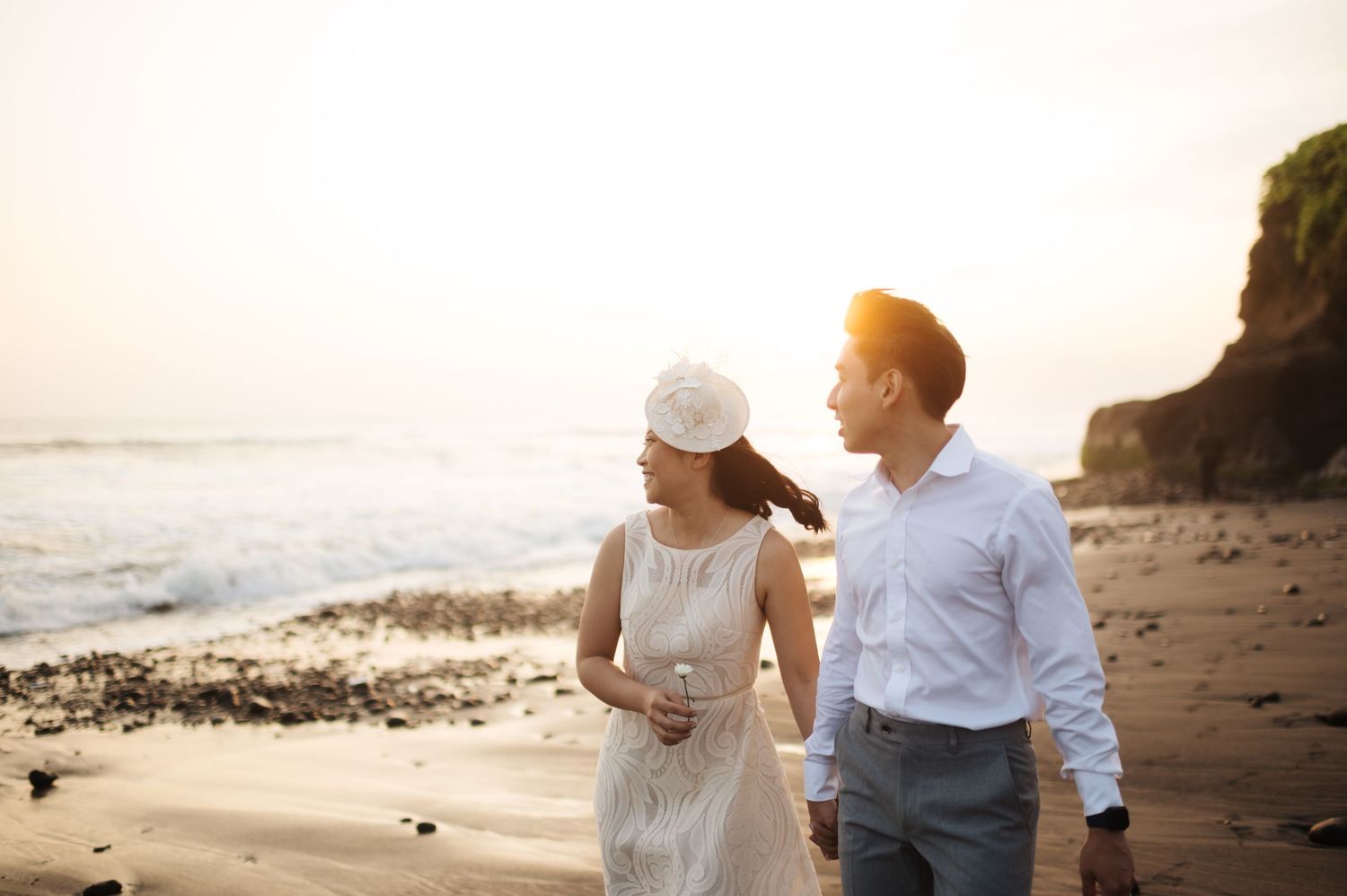 A couple walks hand in hand on a sandy beach at sunset, with ocean waves and rocky cliffs in the background.