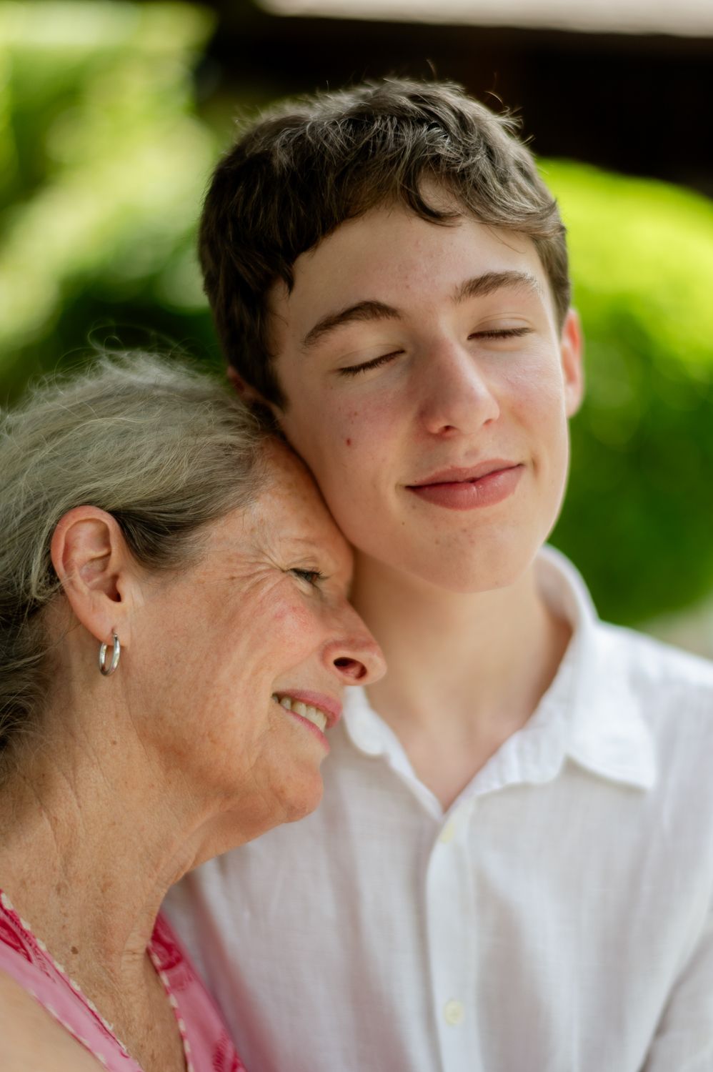 Elderly woman and teenage boy share tender moment, eyes closed, cheek to cheek outdoors.