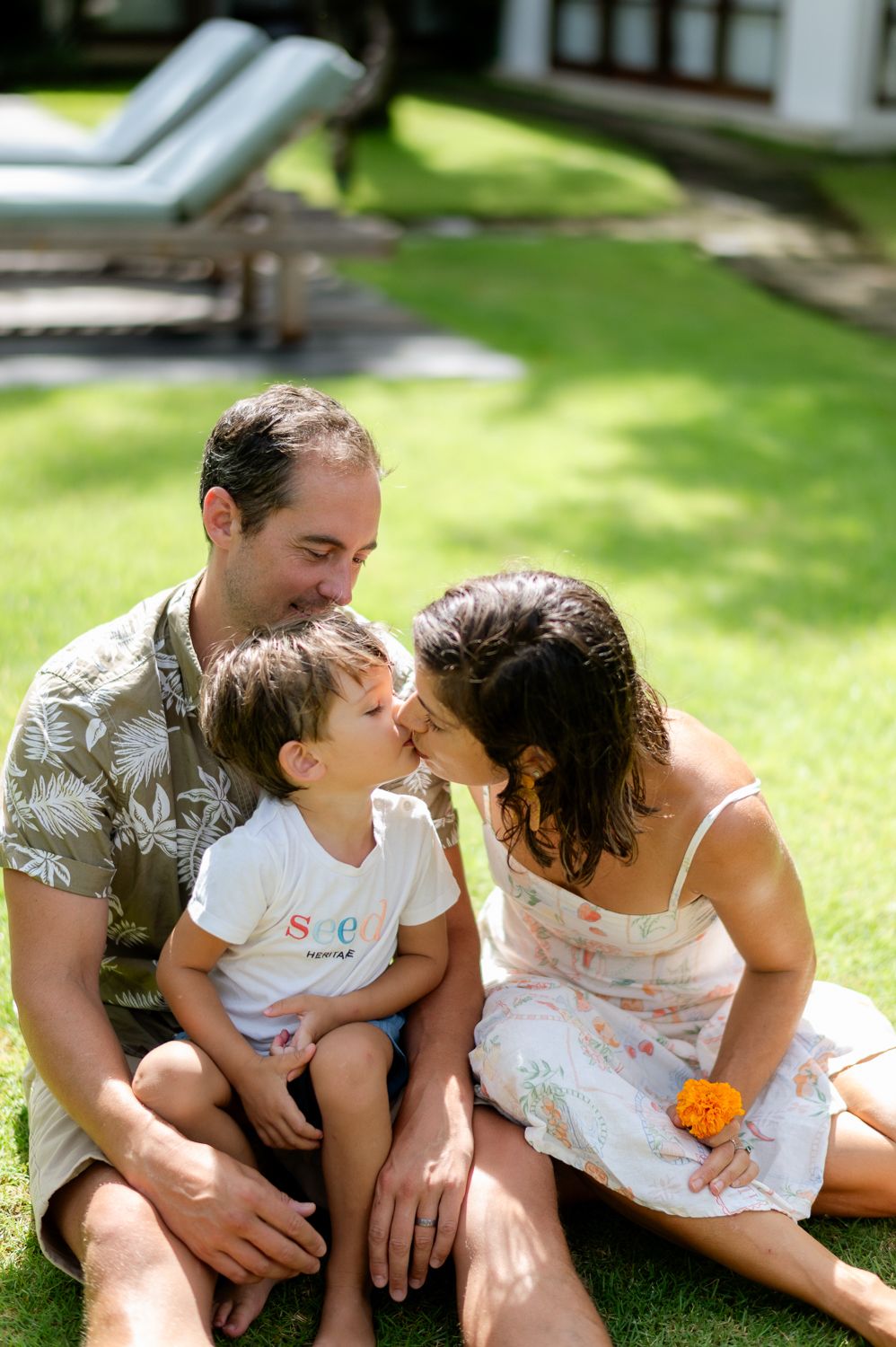 A family of three sits on green grass, parents sharing a kiss with their toddler son outdoors.