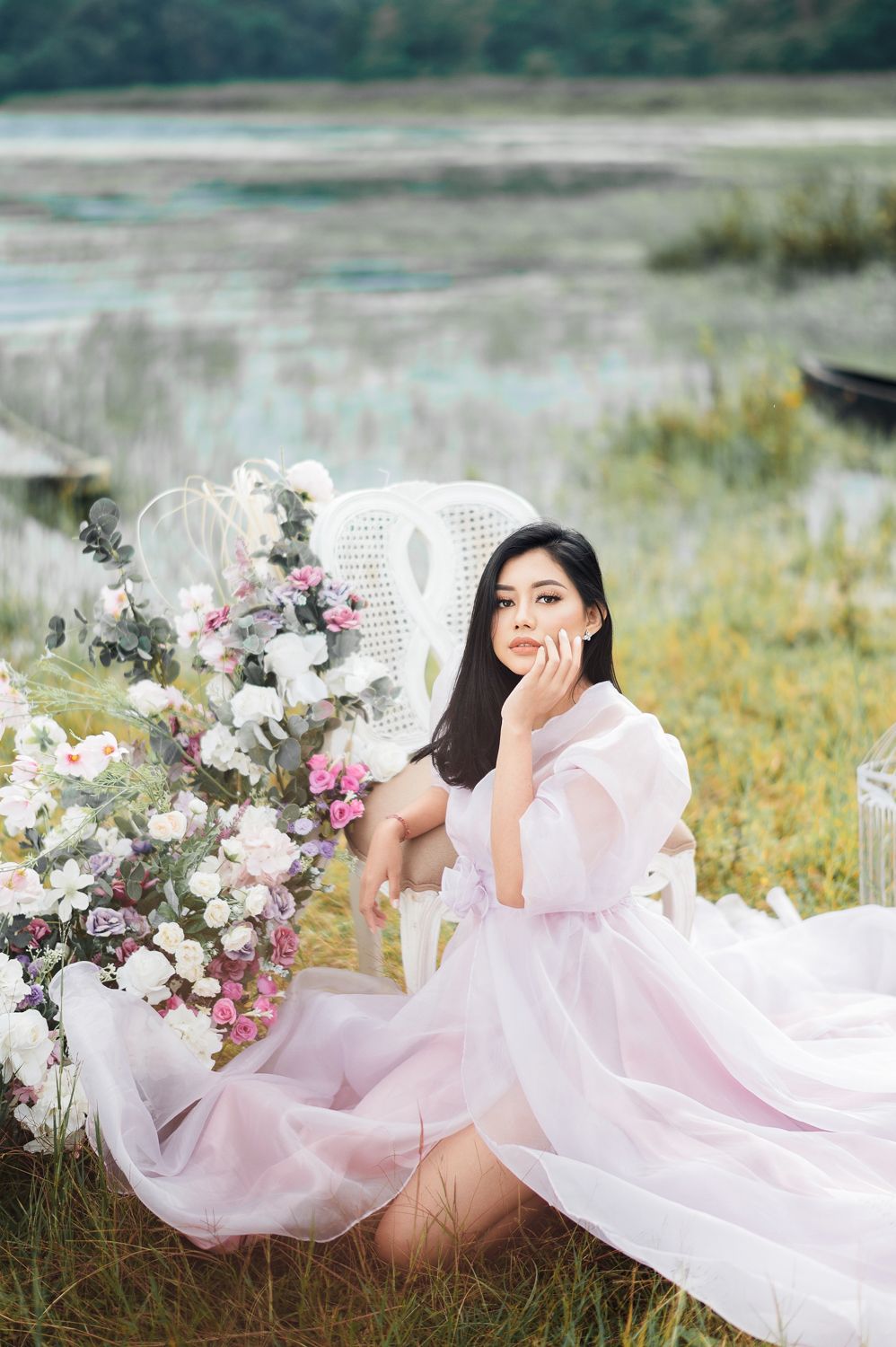 A young woman in a flowing pink tulle gown sits gracefully outdoors near florals and a white chair by a serene lake.
