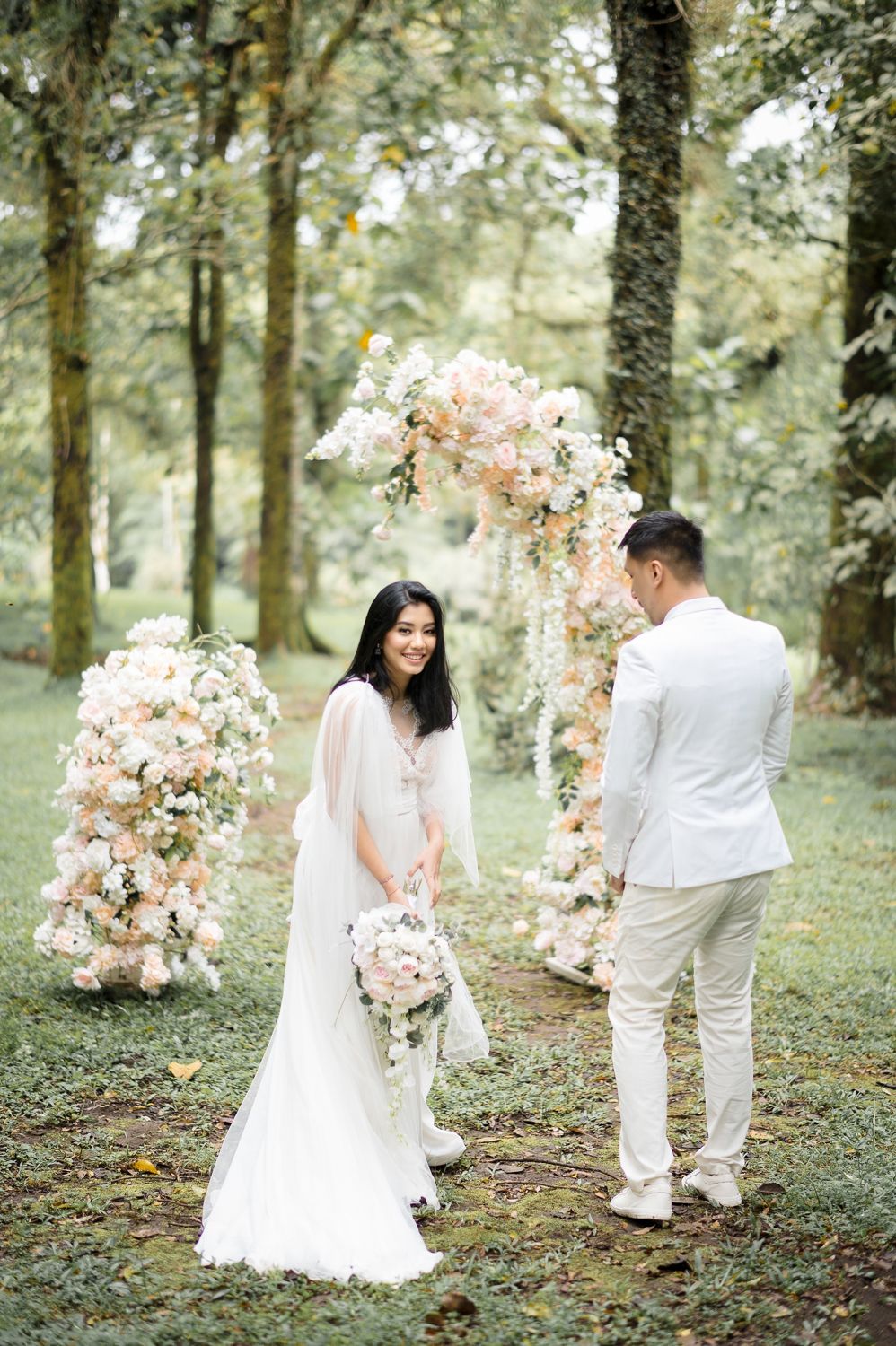 Bride and groom stand beneath a floral arch with blush roses in a lush forest wedding ceremony setting.