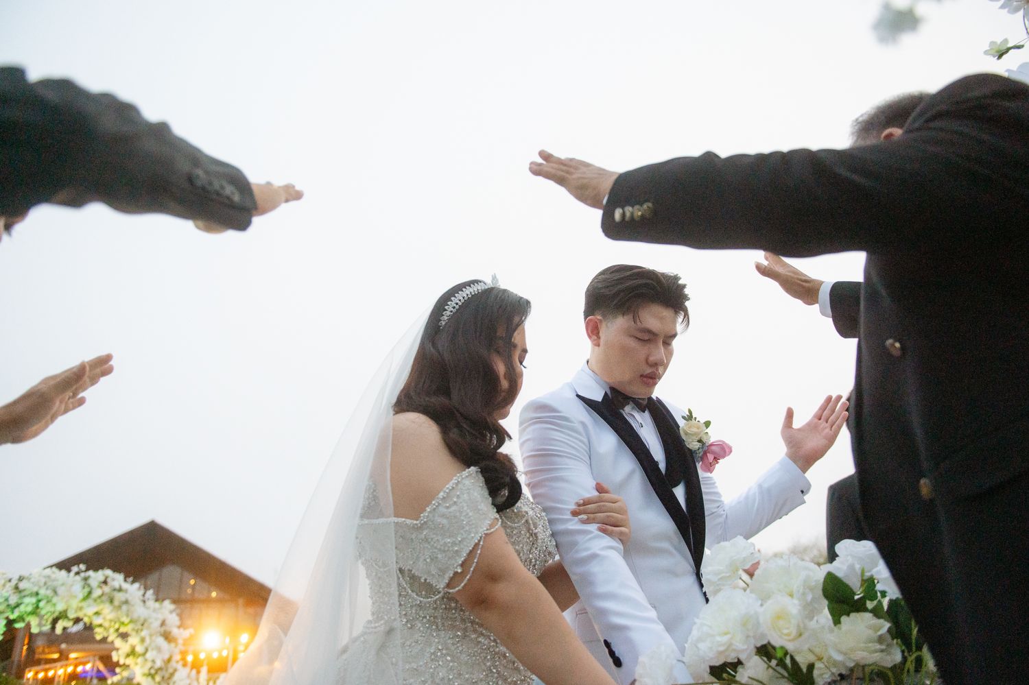 Bride and groom bow during outdoor wedding ceremony while guests extend hands in blessing.