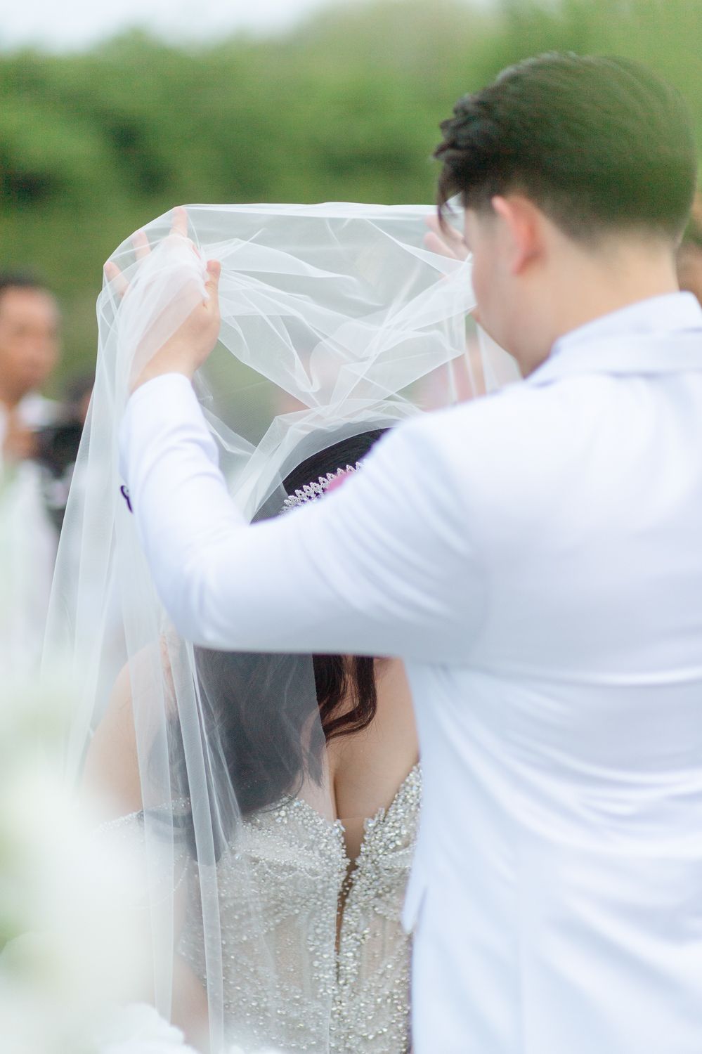 Groom lifting white wedding veil over bride in elegant beaded gown during outdoor ceremony.