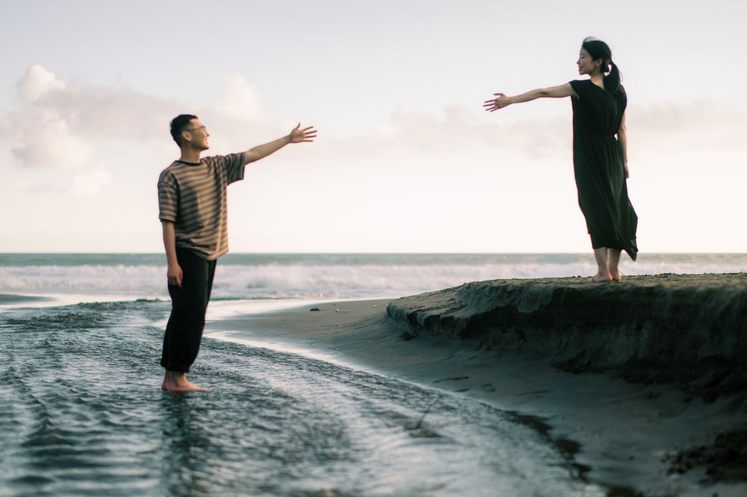 Two people reaching toward each other on a beach at sunset, one standing on sand, the other on a small cliff.