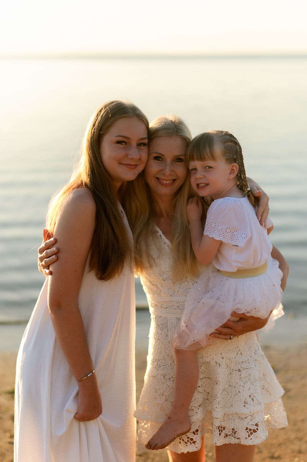 Smiling mother holding young daughter while teenage girl embraces her on a sunlit beach.