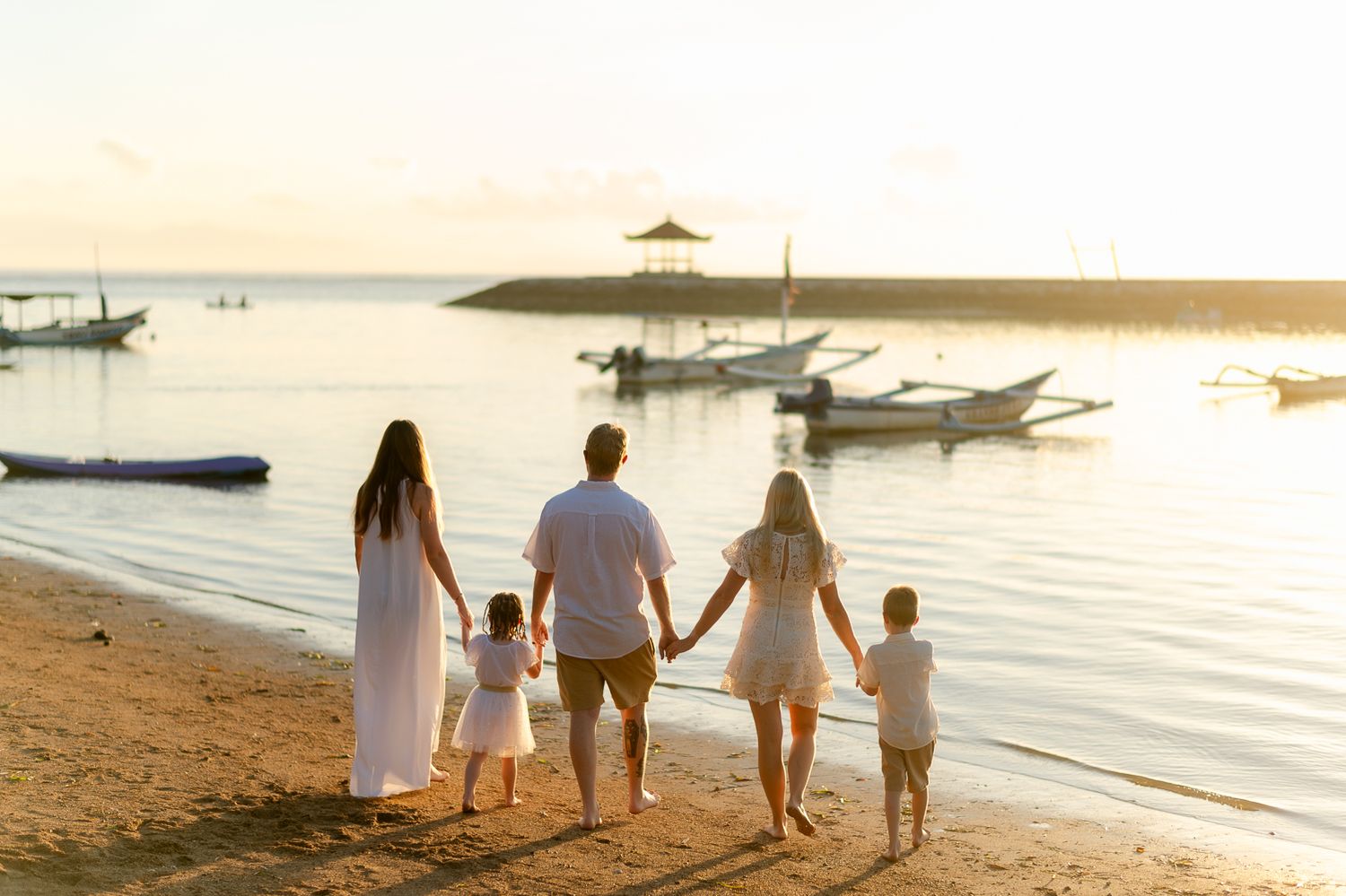 Family of five holding hands walking along a sandy beach at golden sunset with boats in background.