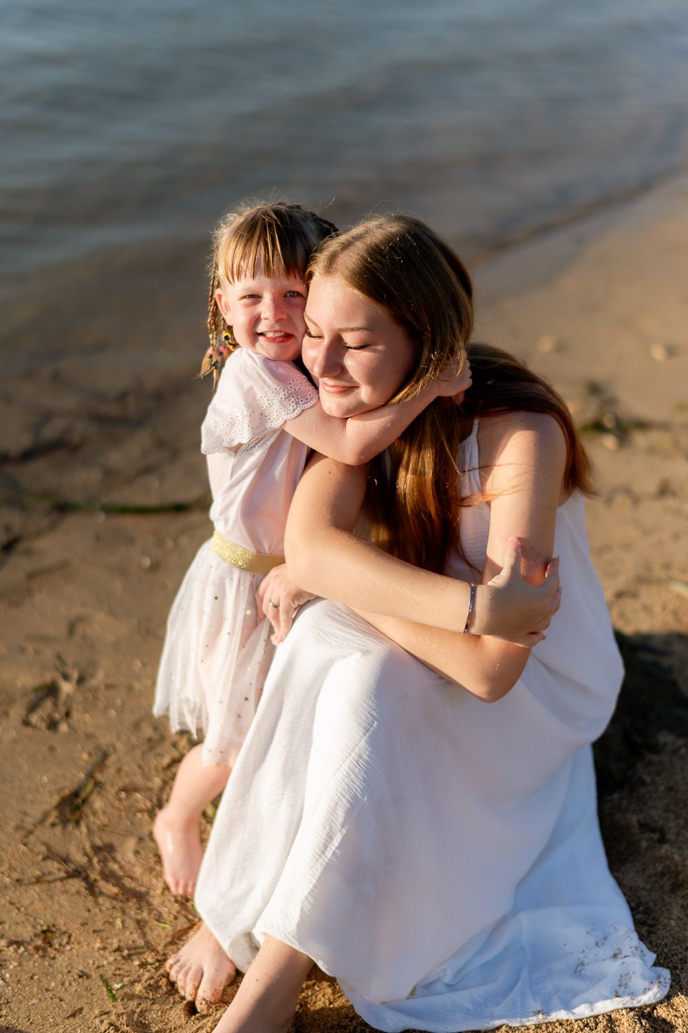 Young girl lovingly hugs older sister in white dress on sandy beach at sunset.