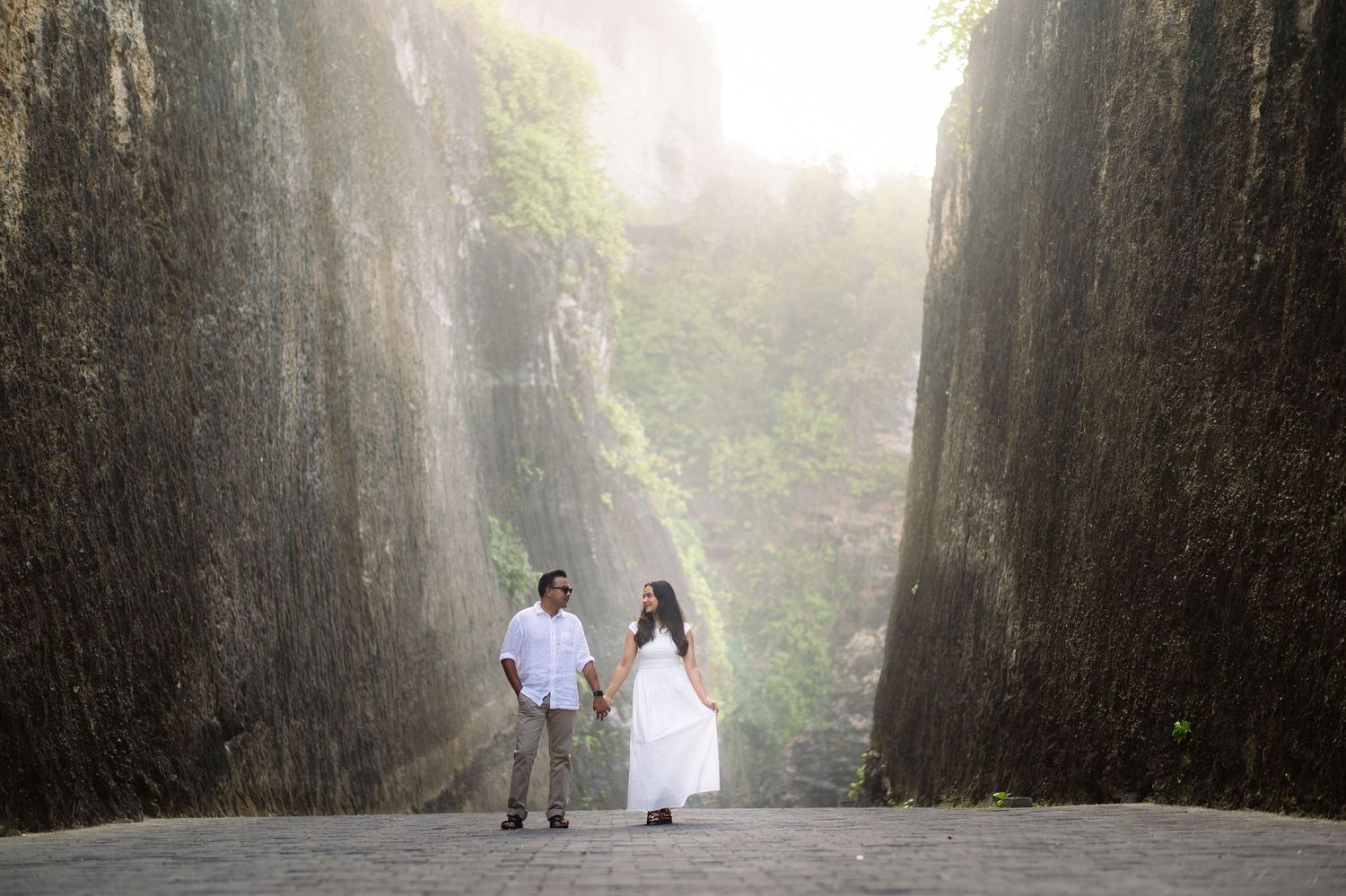 Couple in white holding hands between towering rock walls with misty light, romantic outdoor photo shoot.