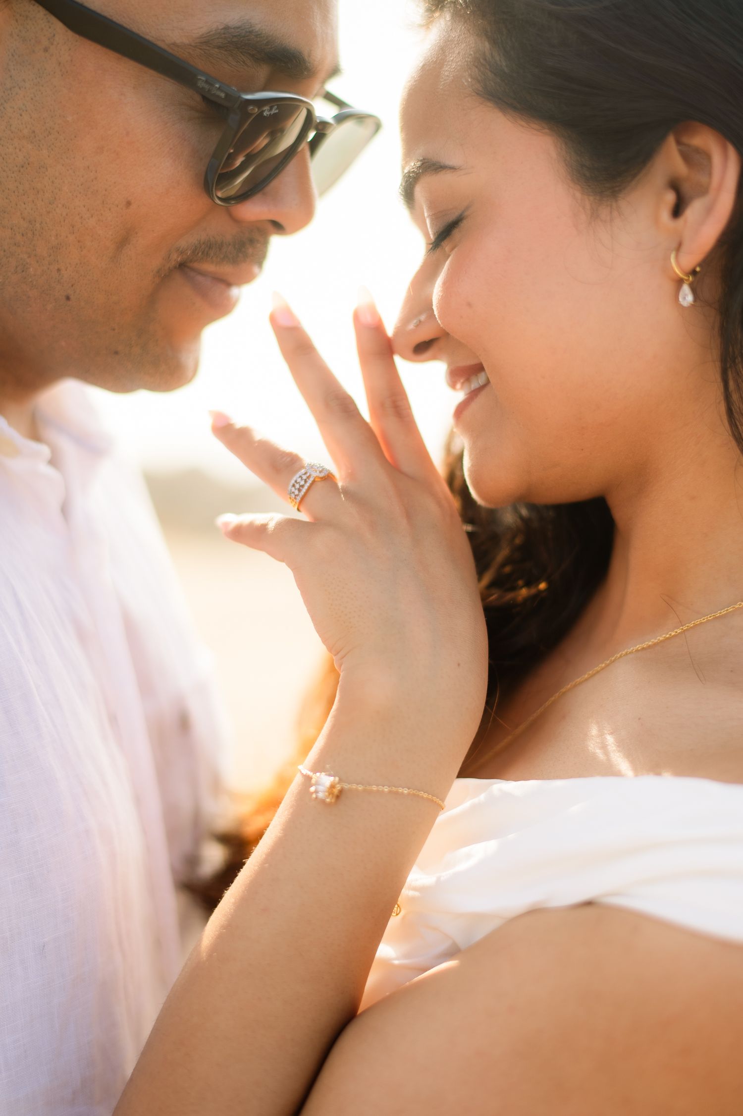 Couple sharing a romantic moment, woman smiling with engagement ring visible, golden backlit glow, delicate jewelry.