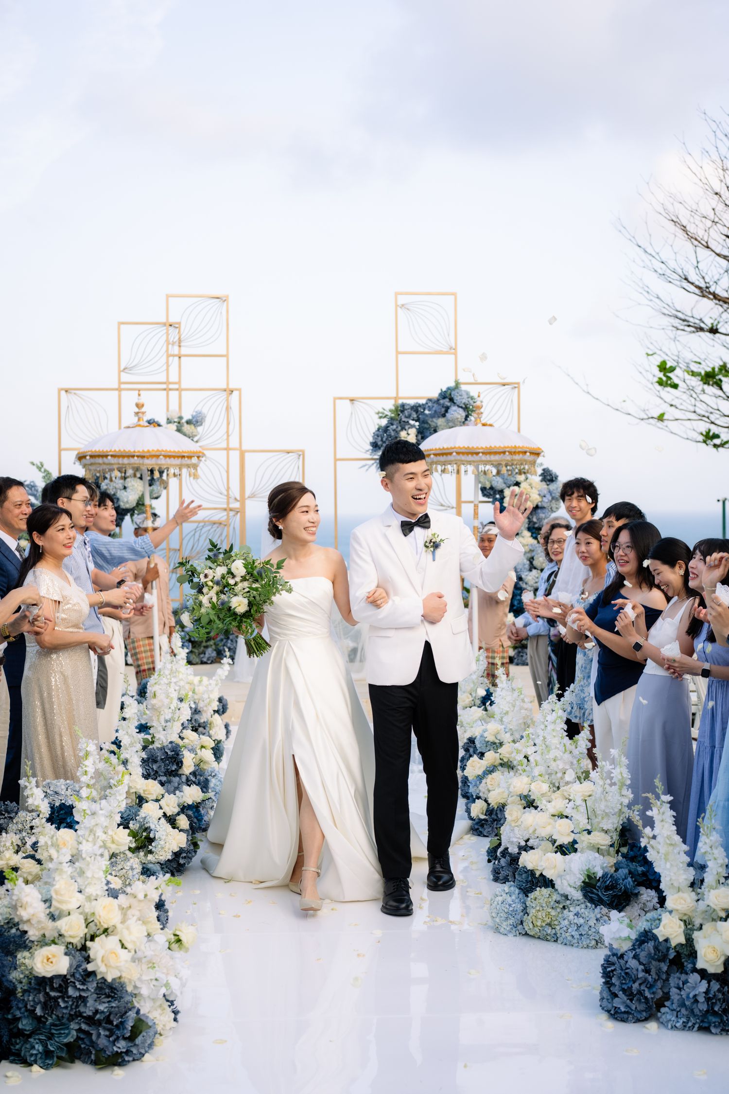 Bride and groom walk hand in hand down an aisle lined with blue and white flowers as cheering guests celebrate at an oceanview wedding.