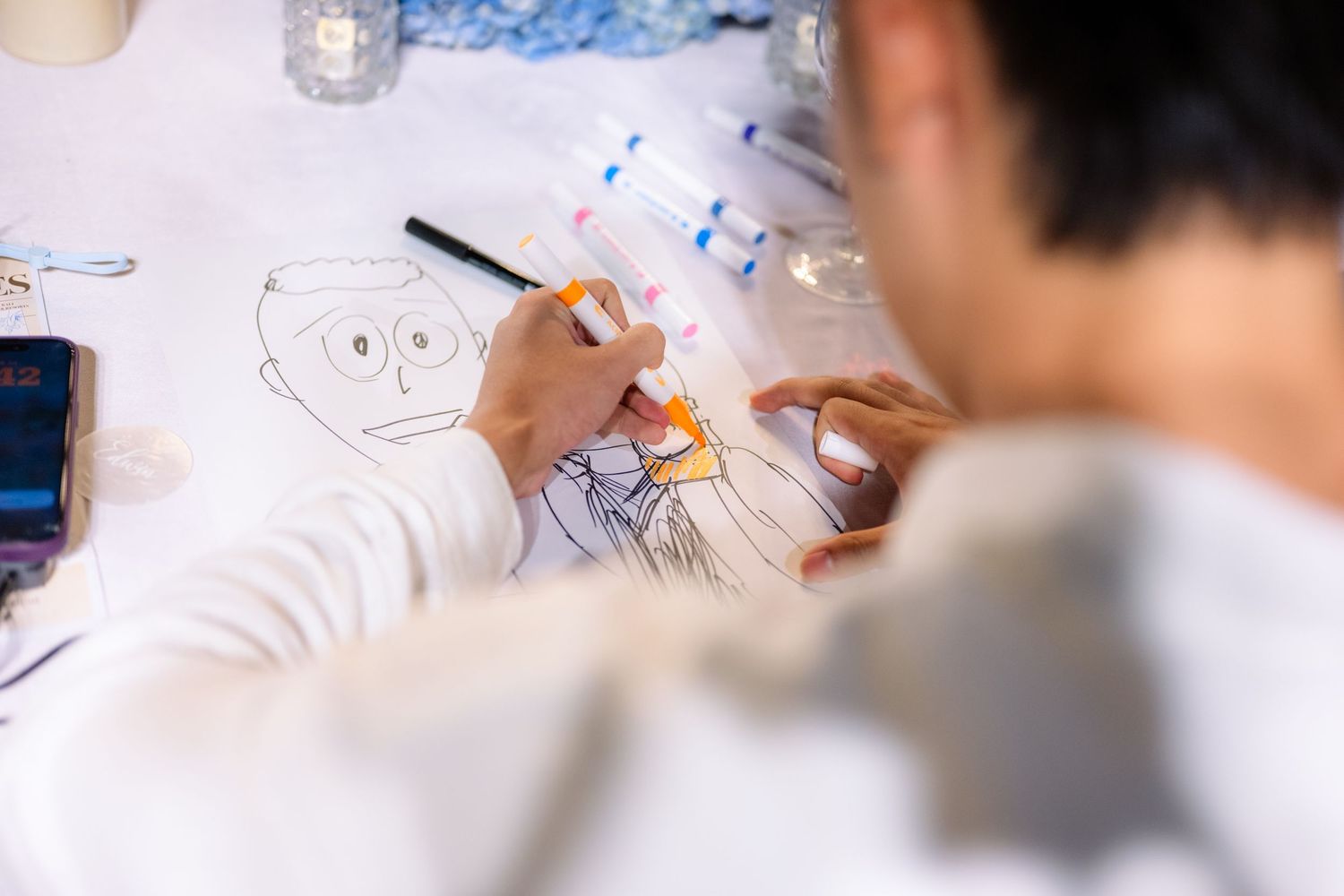 Person in a white sweatshirt coloring a cartoon face drawing with markers on a paper-covered table, with a smartphone nearby.