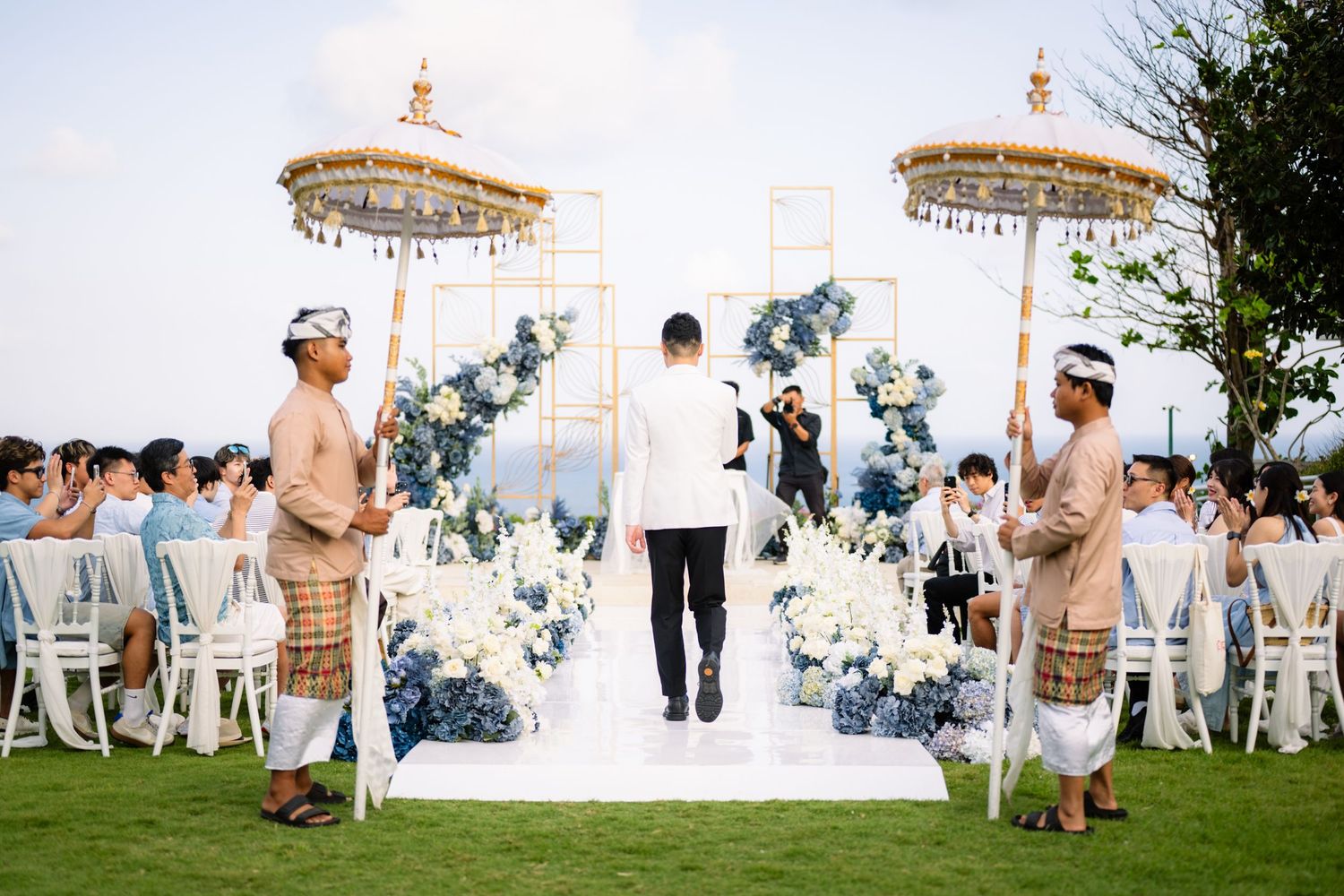 Groom stands at a Balinese outdoor wedding ceremony with blue and white floral arrangements, golden umbrellas, and guests seated on a lawn.