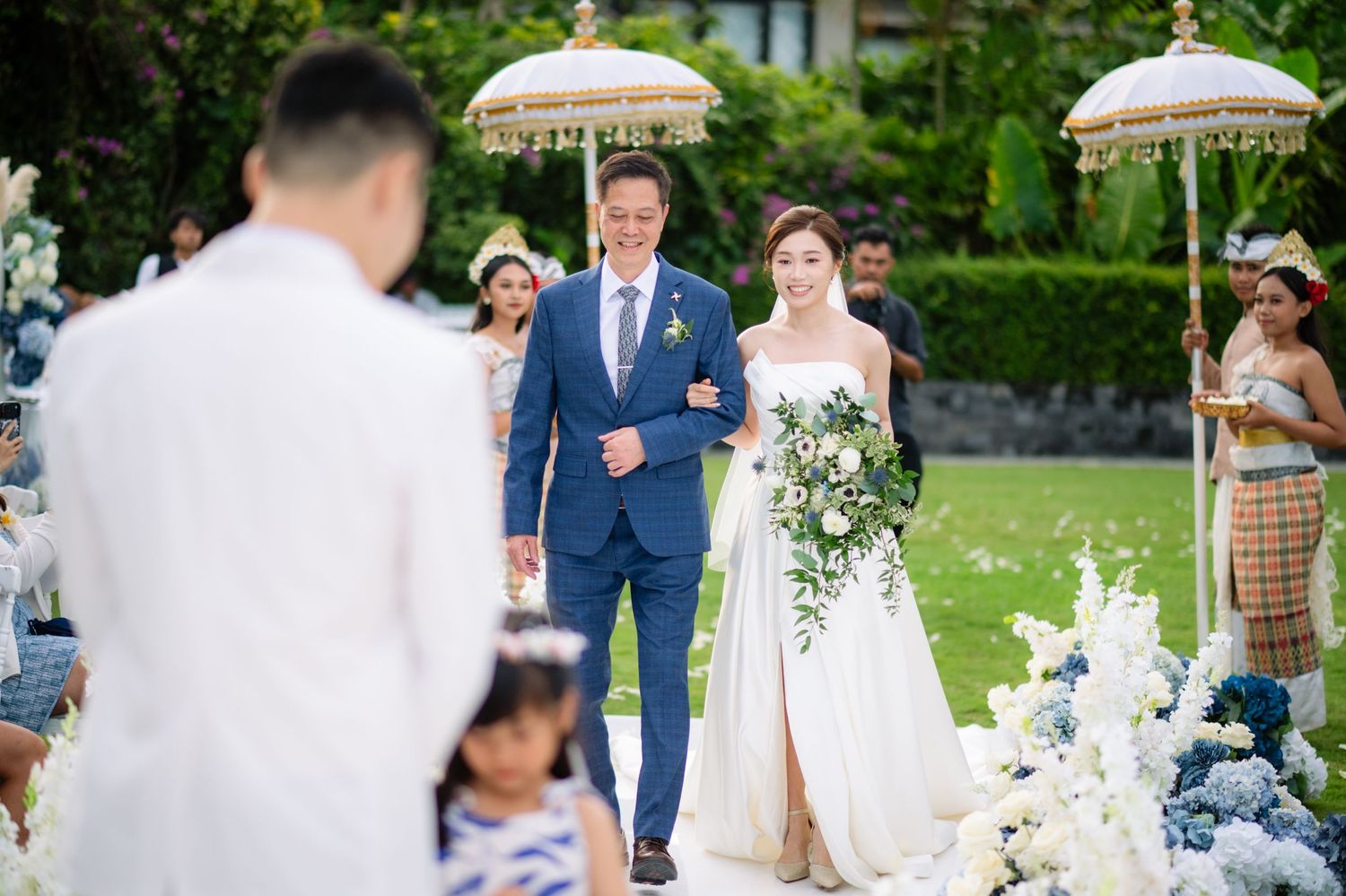 Bride in a white gown holding a bouquet walks arm in arm with the groom in a blue suit down an outdoor garden wedding aisle.