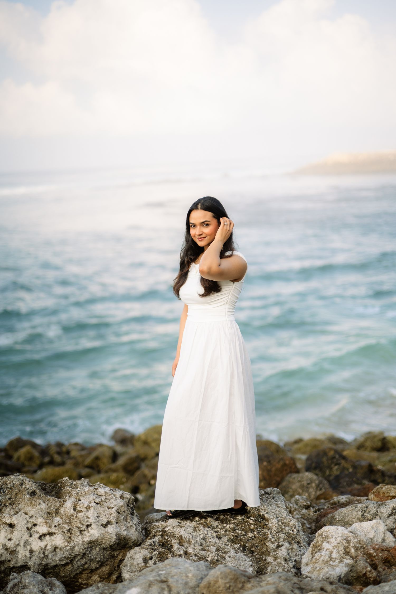 Woman in white maxi dress standing on rocky shoreline with turquoise ocean waves in the background.