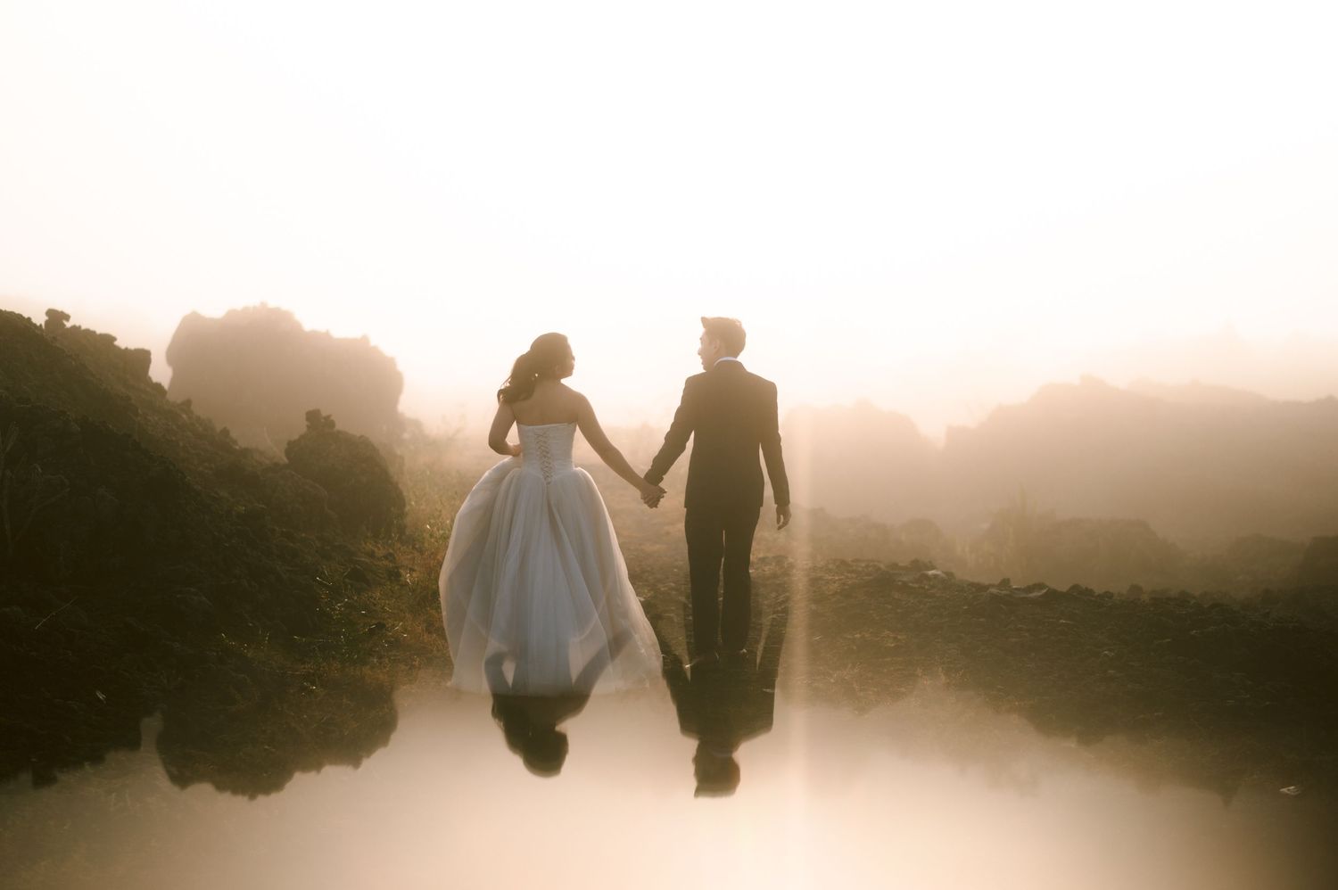 Bride and groom holding hands walking into misty fog, reflected in still water during golden hour wedding photography.