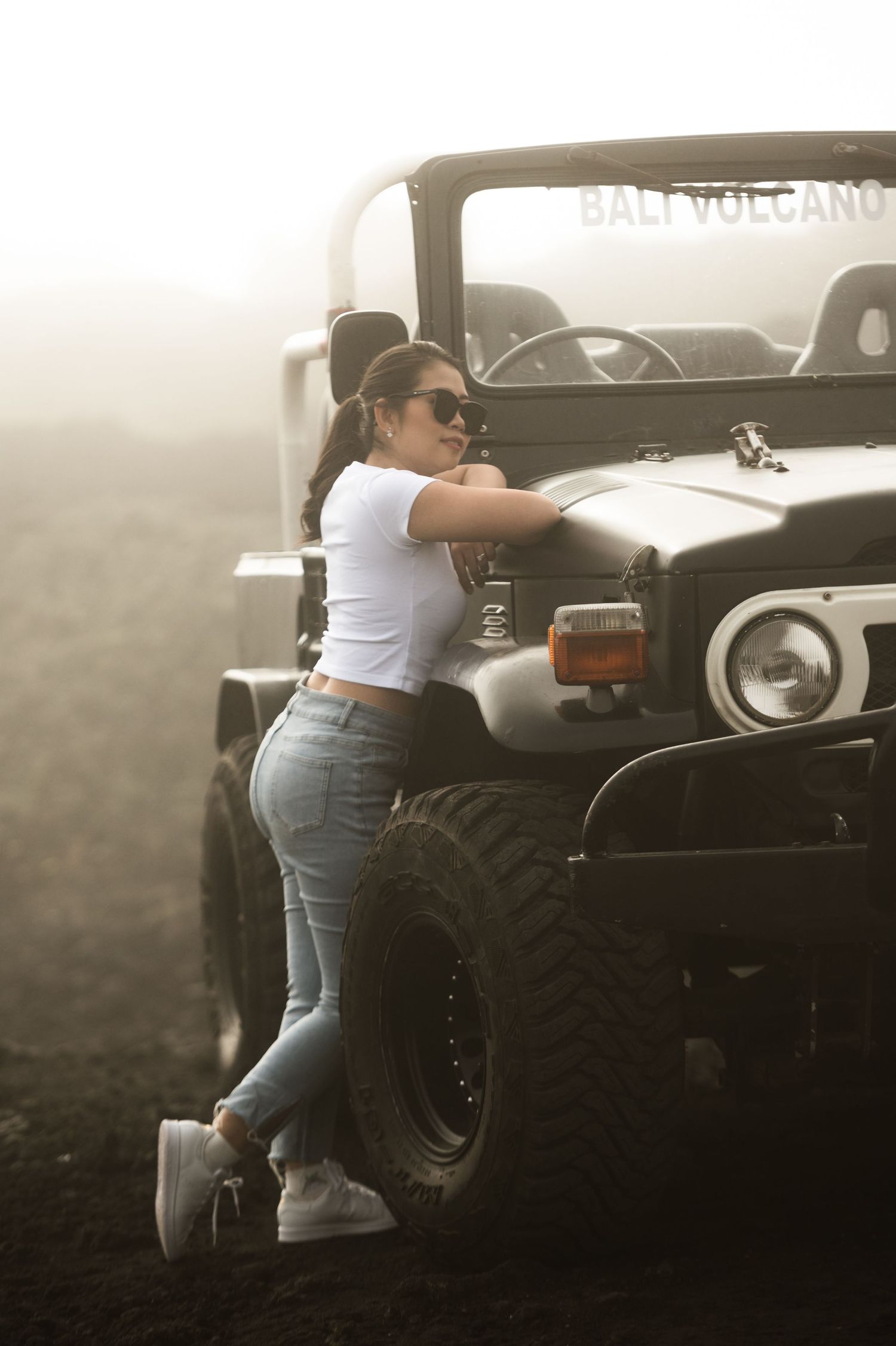 Woman in white crop top and jeans leaning on a jeep at Bali Volcano, wearing sunglasses in misty conditions.