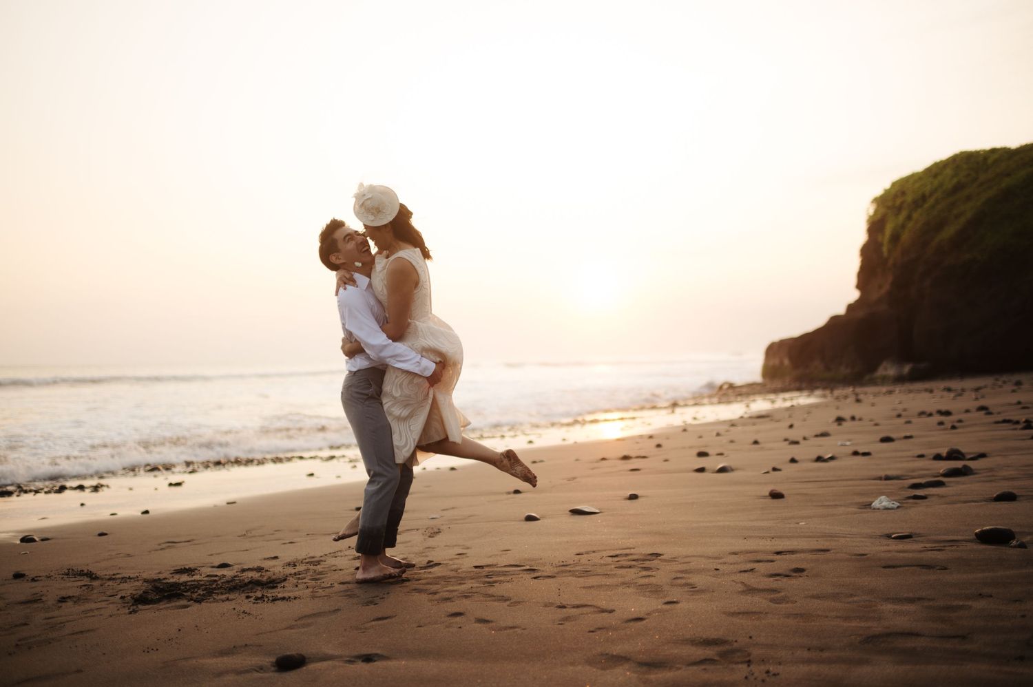 A couple shares a romantic kiss on a sandy beach at sunset, with the groom lifting the bride in her white dress.