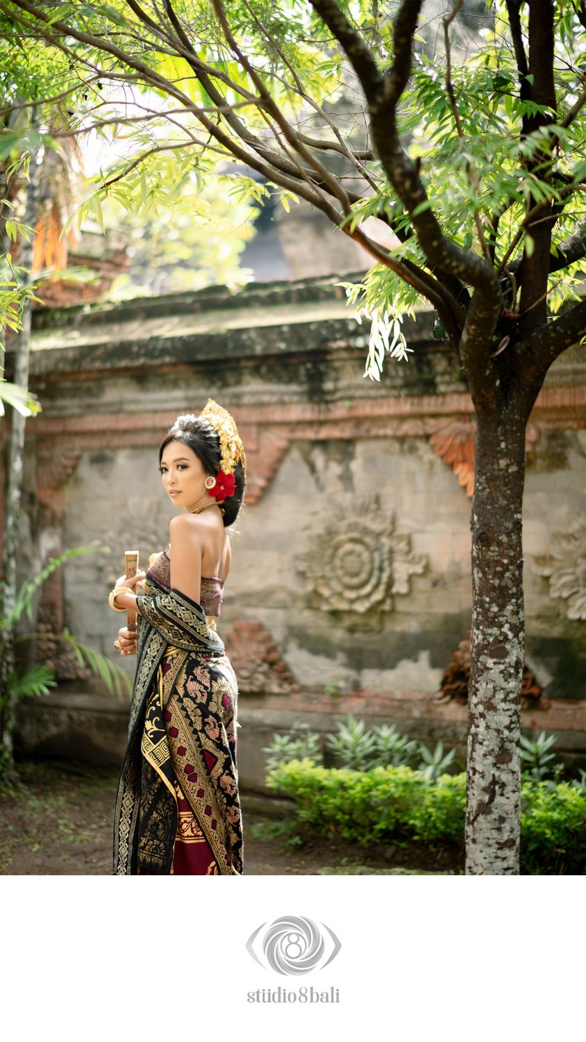 Balinese woman in traditional attire poses gracefully near ancient stone temple wall and tree in Bali, Indonesia.