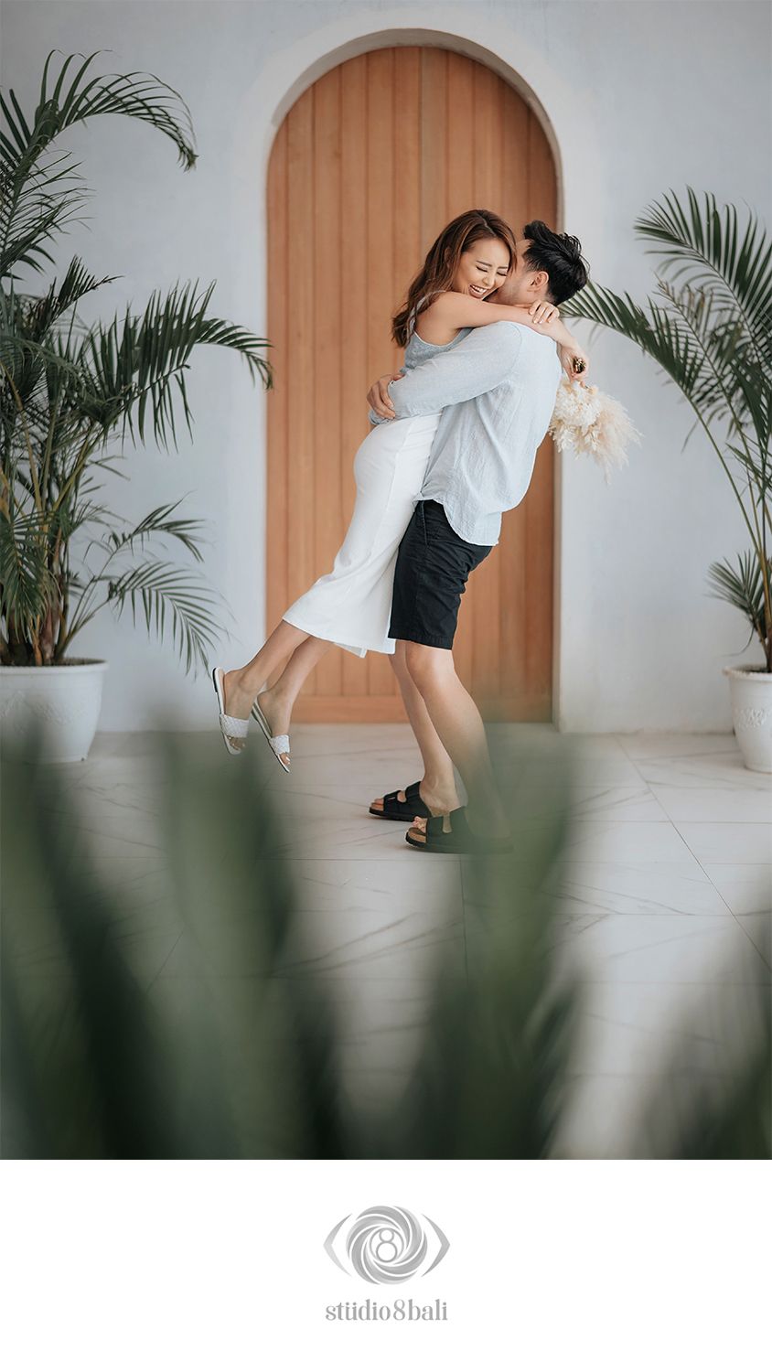 Couple embracing in a romantic lift pose before an arched wooden door, surrounded by tropical plants in a bright studio.