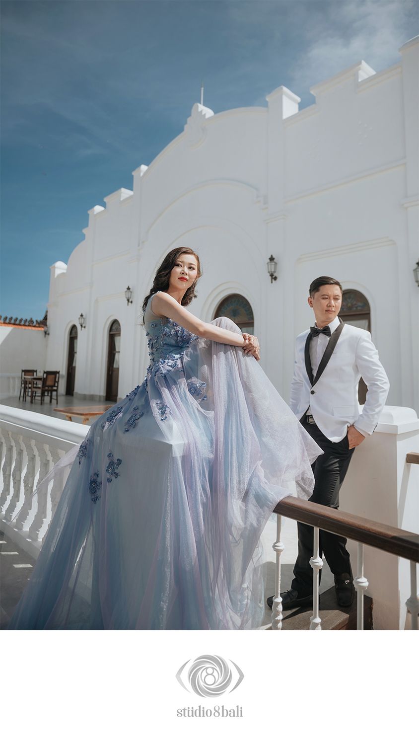 Bride in flowing blue floral gown poses with groom in white tuxedo on a sunlit white colonial terrace.
