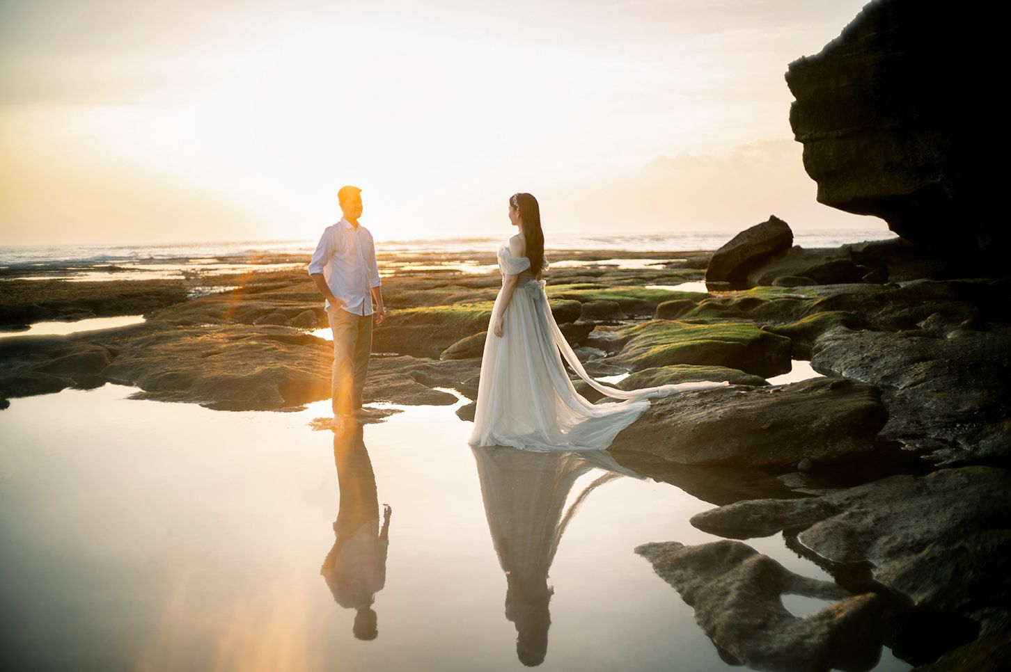 Couple in wedding attire standing on rocky shore at sunset with reflections in tide pools.