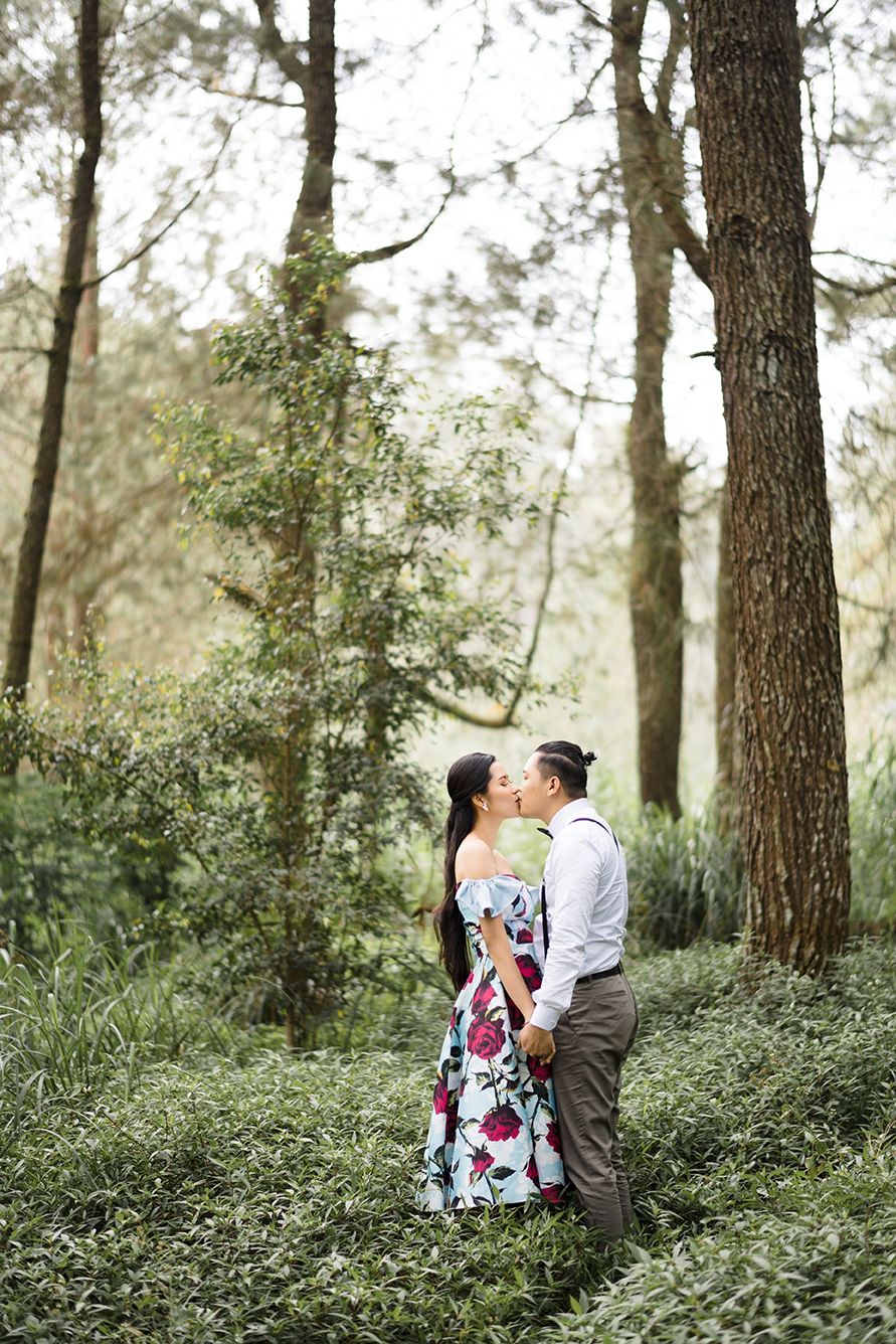 Couple sharing a romantic kiss in a lush green forest, woman wearing a floral off-shoulder dress, man in white shirt.