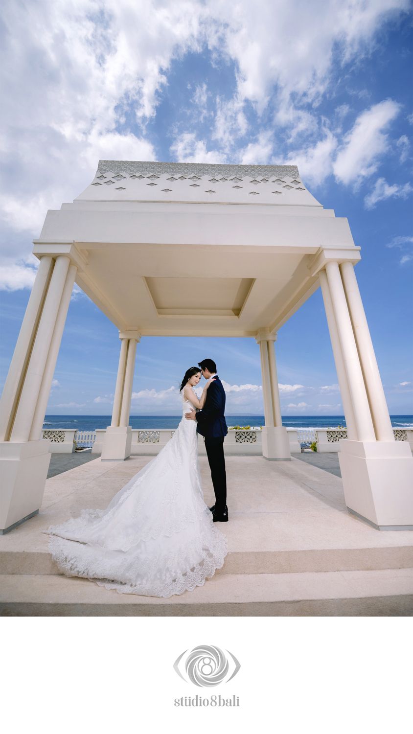 Bride and groom embrace under elegant white oceanfront pavilion with blue sky backdrop at a destination wedding in Bali.