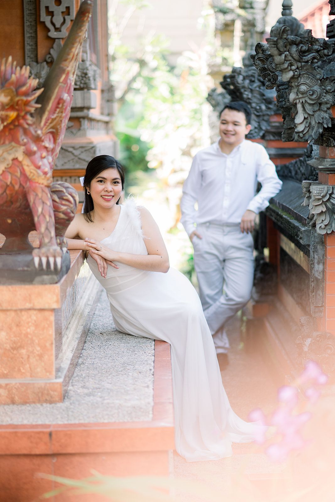 Couple in white outfits posing at a Balinese temple, woman in elegant dress seated on steps, man standing behind.