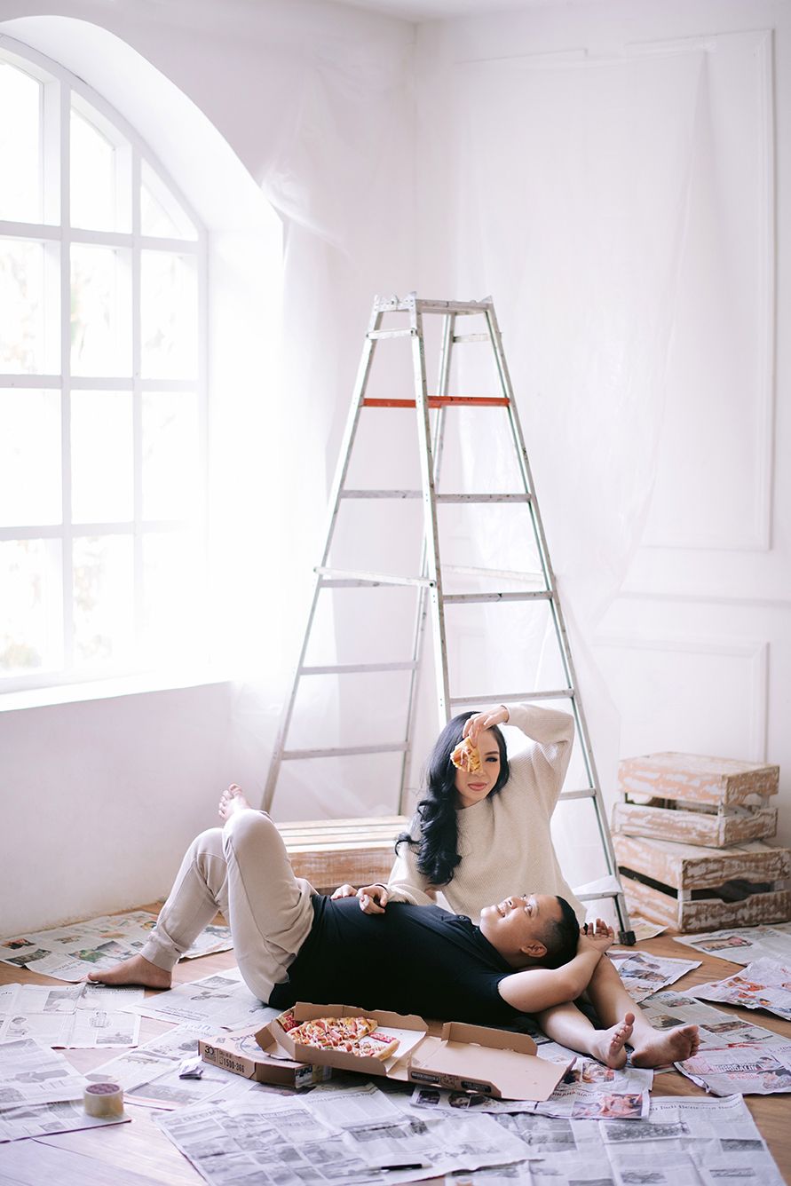 Couple relaxing on newspaper-covered floor near ladder during home renovation, sharing pizza in bright white room.