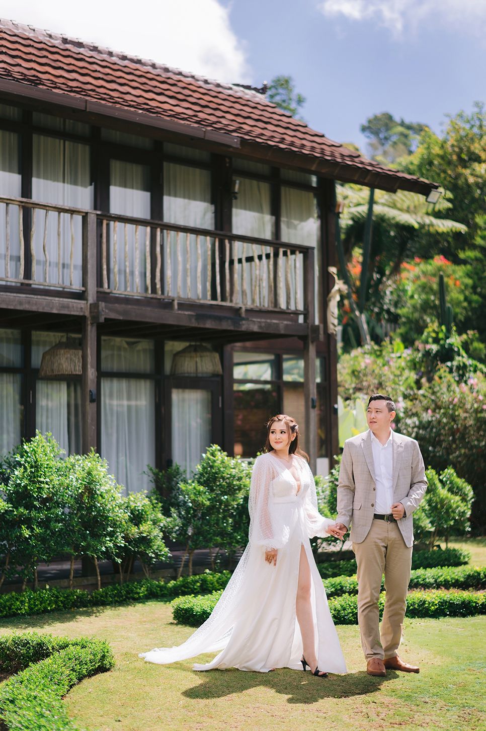 Couple in wedding attire posing in lush garden before elegant two-story colonial building with wooden balcony.