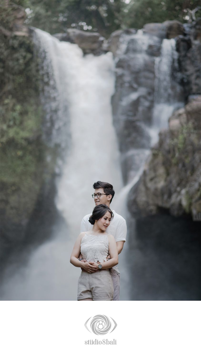 Couple embracing romantically in front of a stunning waterfall backdrop in Bali, wearing matching beige outfits.
